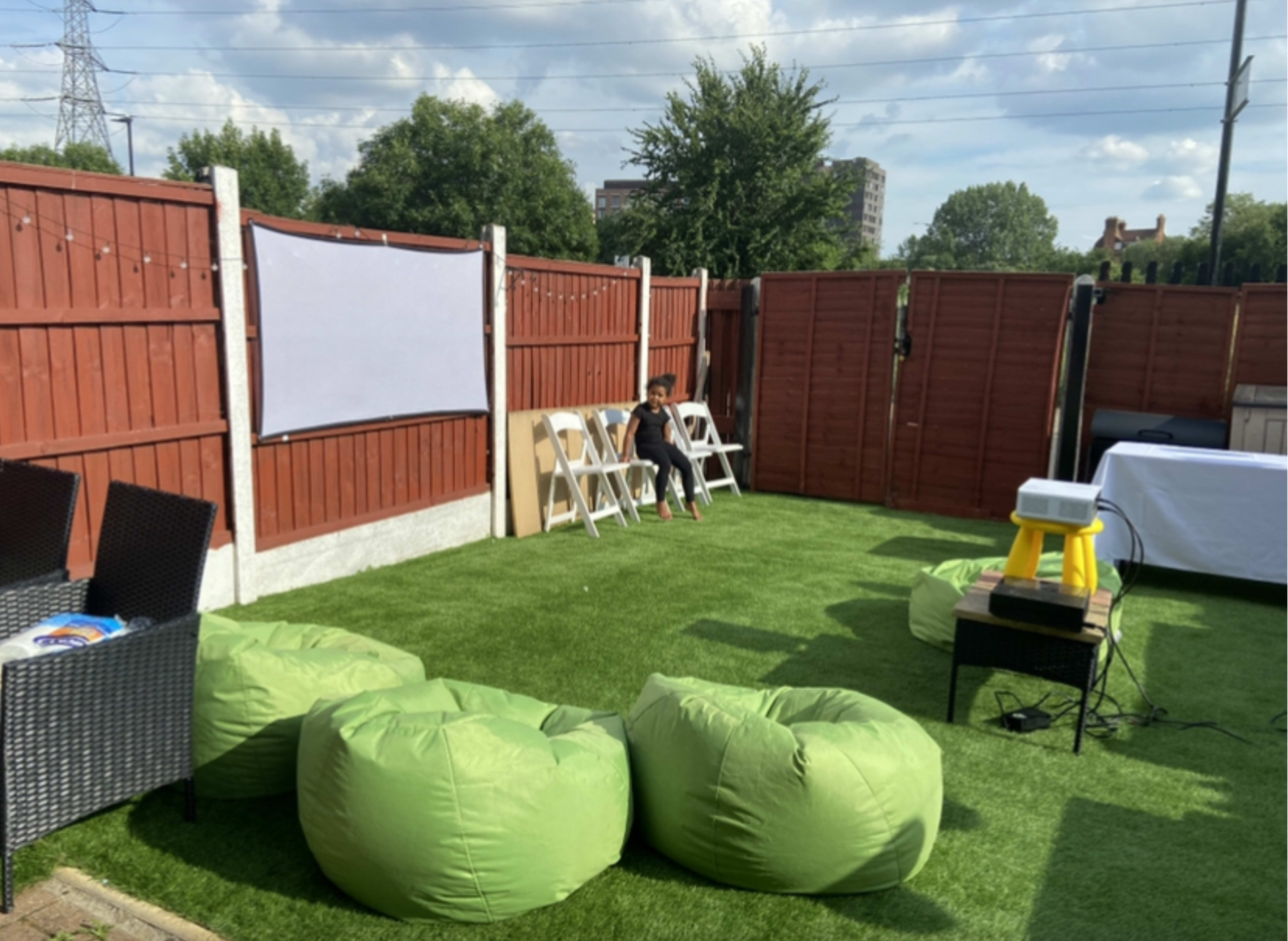 A person sits on a chair in a fenced backyard decorated with green bean bag chairs and a white projector screen.