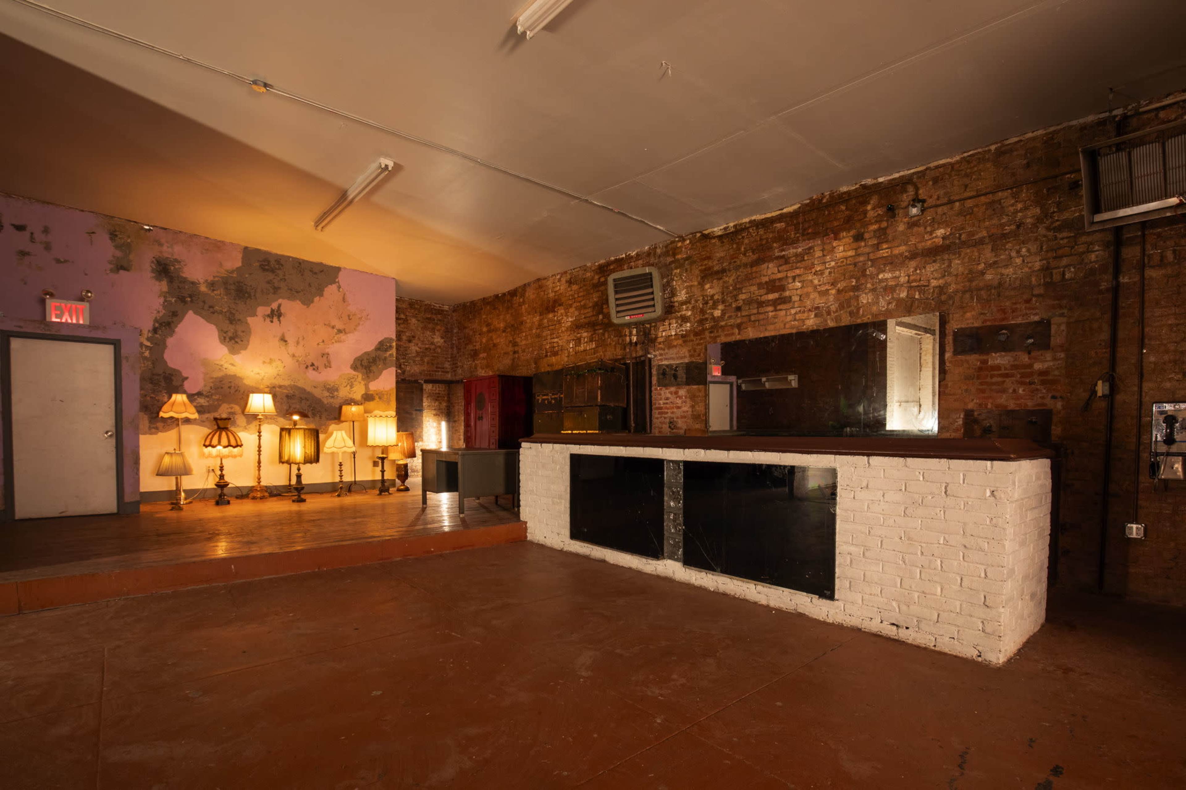 The interior of a room features a bar counter with a black and white design, vintage lamps on the floor, and exposed brick walls with a partially painted section.