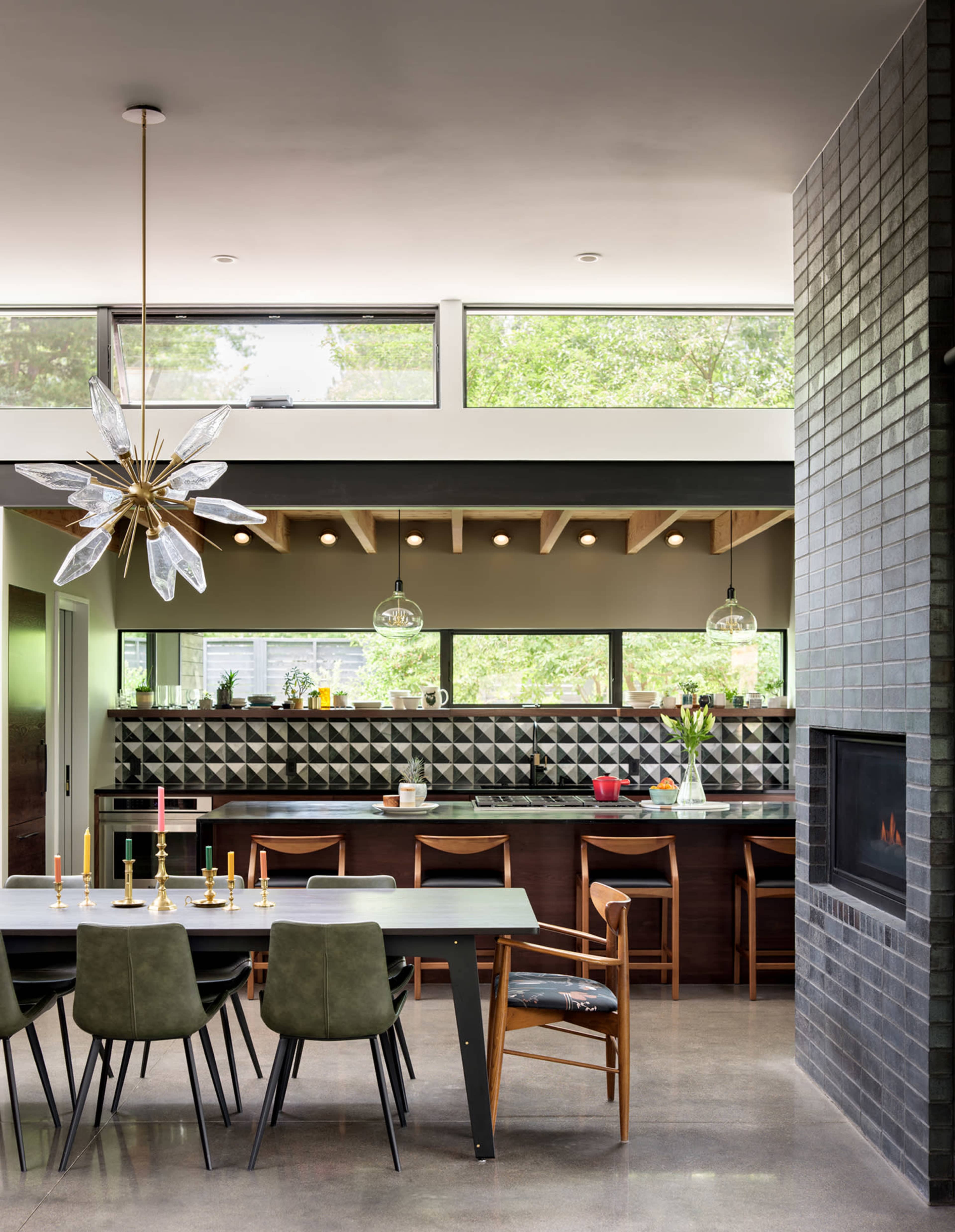 A modern kitchen and dining area features a long table with chairs, a sleek counter with geometric tile backsplash, and large windows letting in natural light.