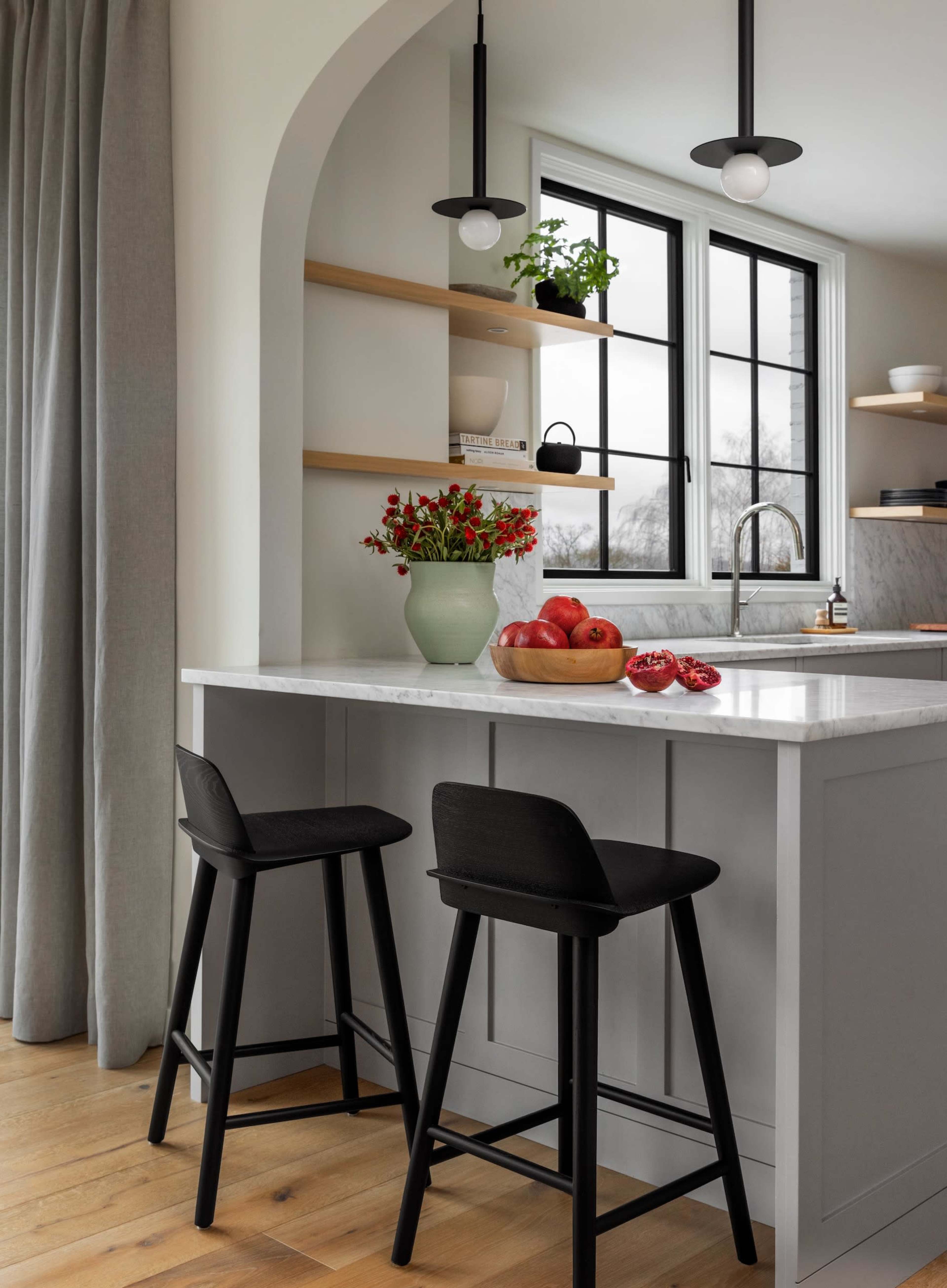 A modern kitchen features a white countertop island with black stools, a vase of flowers, and a bowl of red apples, along with shelves and large windows.