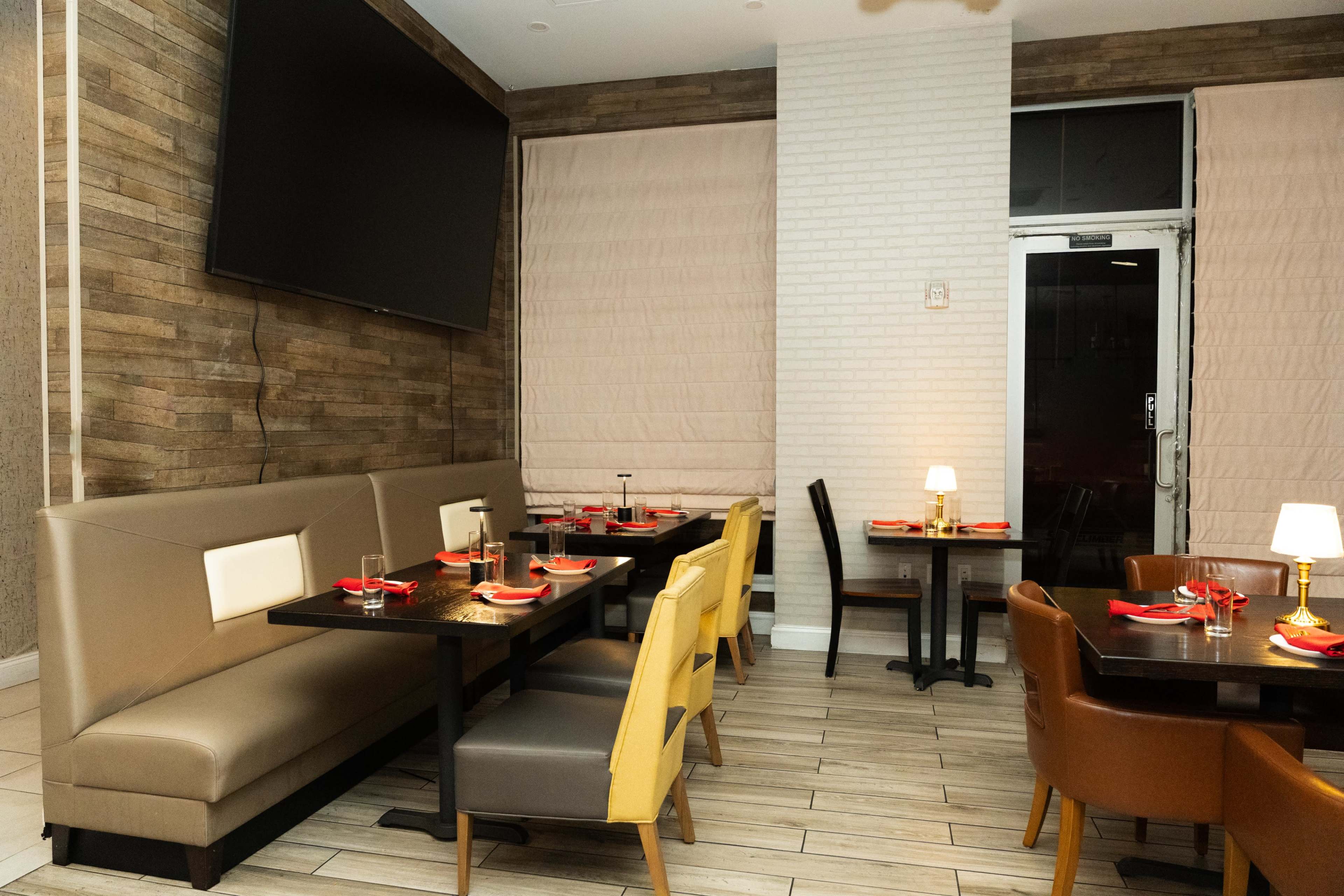 The image shows a modern restaurant interior with a large television mounted on the wall, seating areas with tables set for dining, and large windows covered by shades.