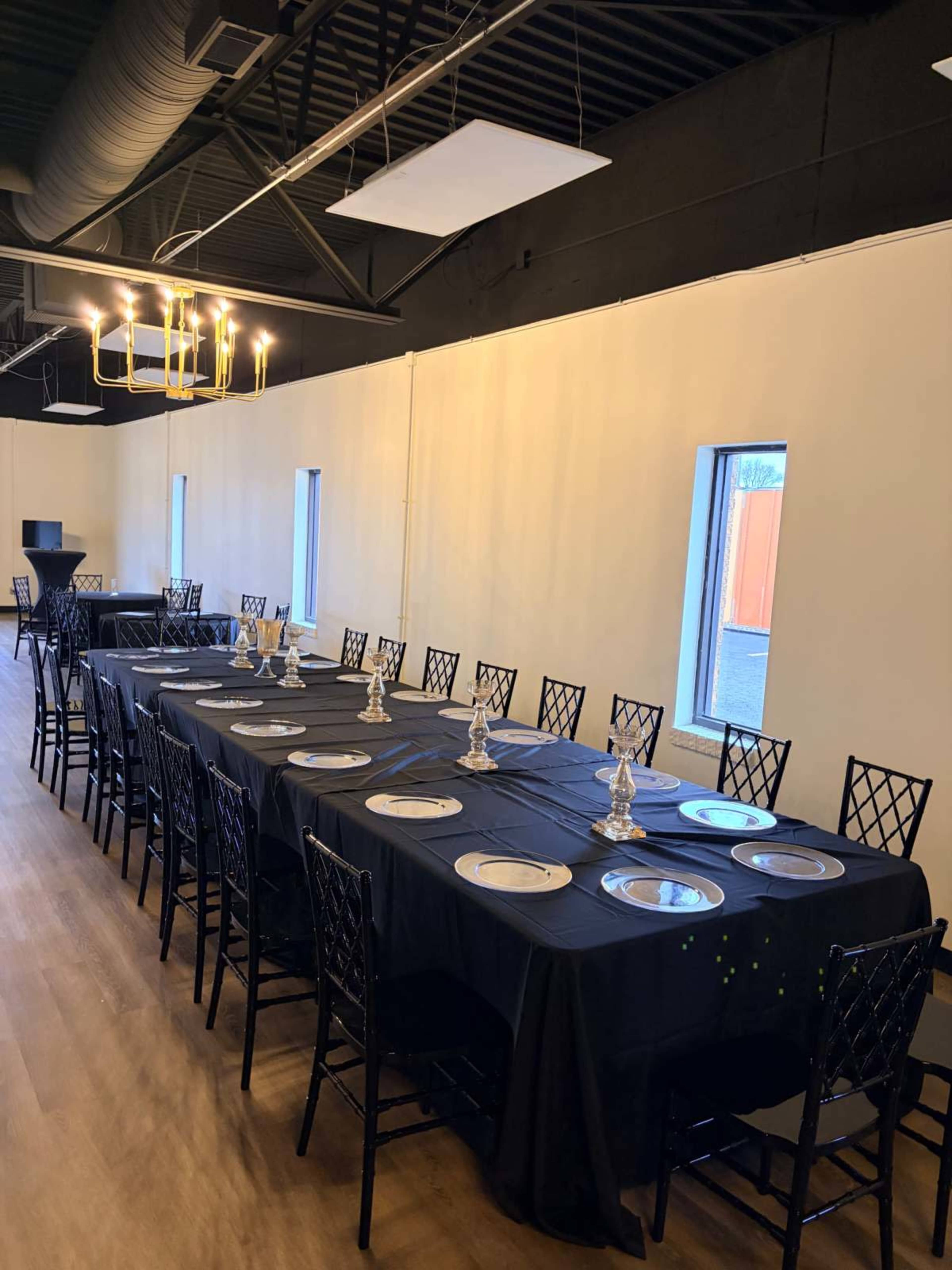 A long dining table is set with black tablecloths and plates, surrounded by black chairs, in a room with white walls and windows.