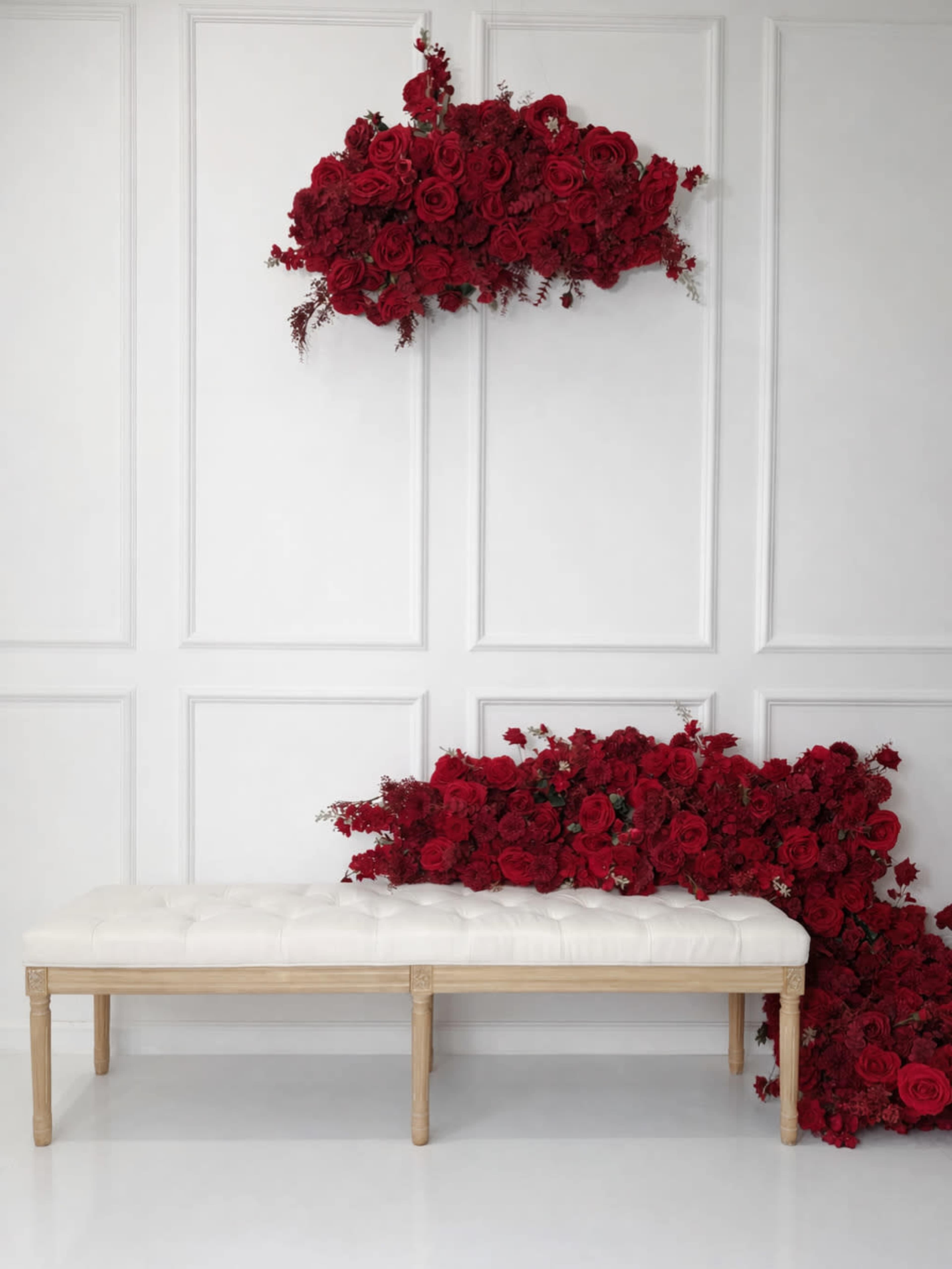 A white bench sits against a wall adorned with a large arrangement of red roses both above and spilling onto the floor.