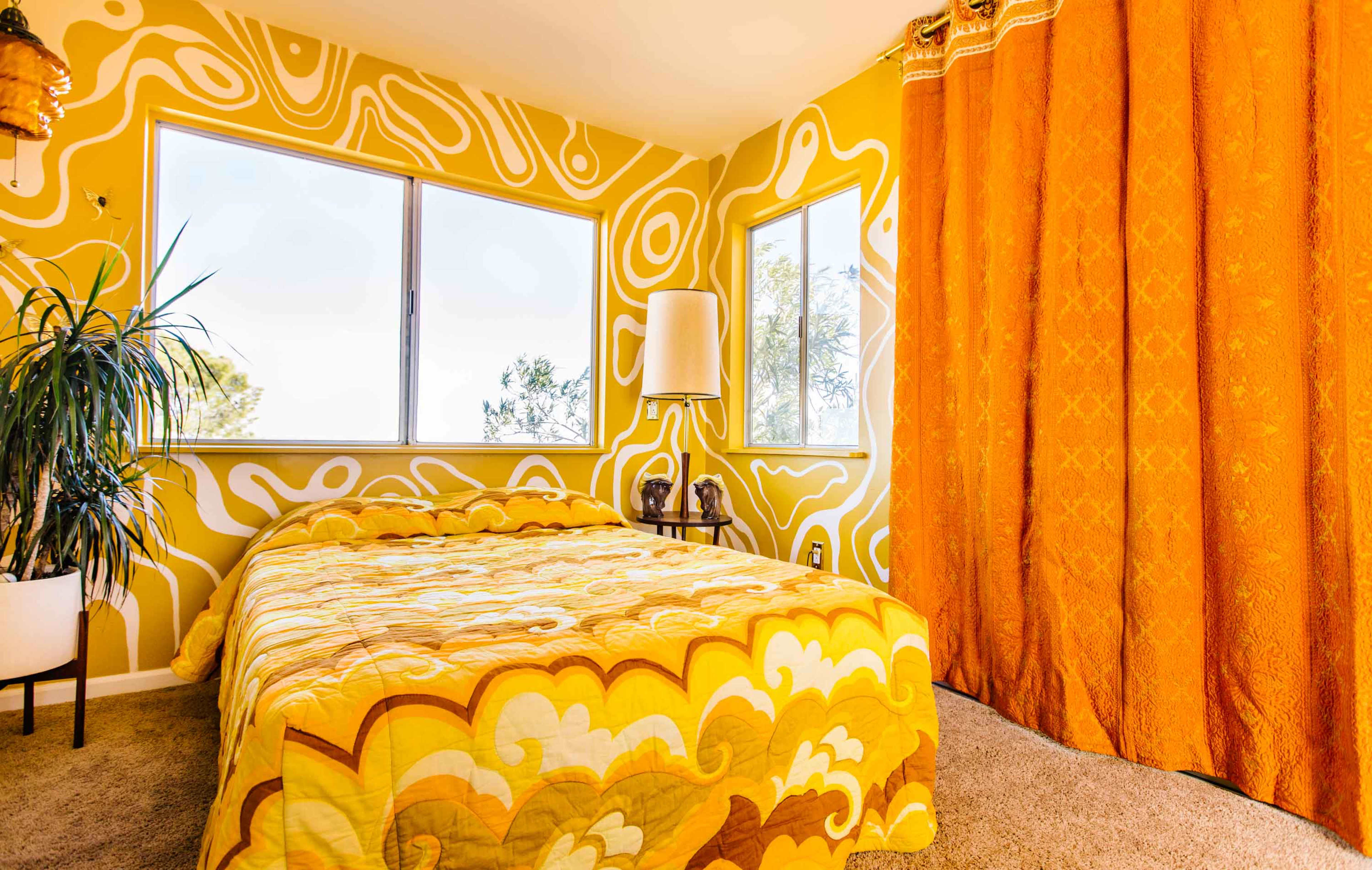 A bedroom with a yellow patterned wall, a bed covered in a similar color scheme, a floor lamp, and orange curtains.