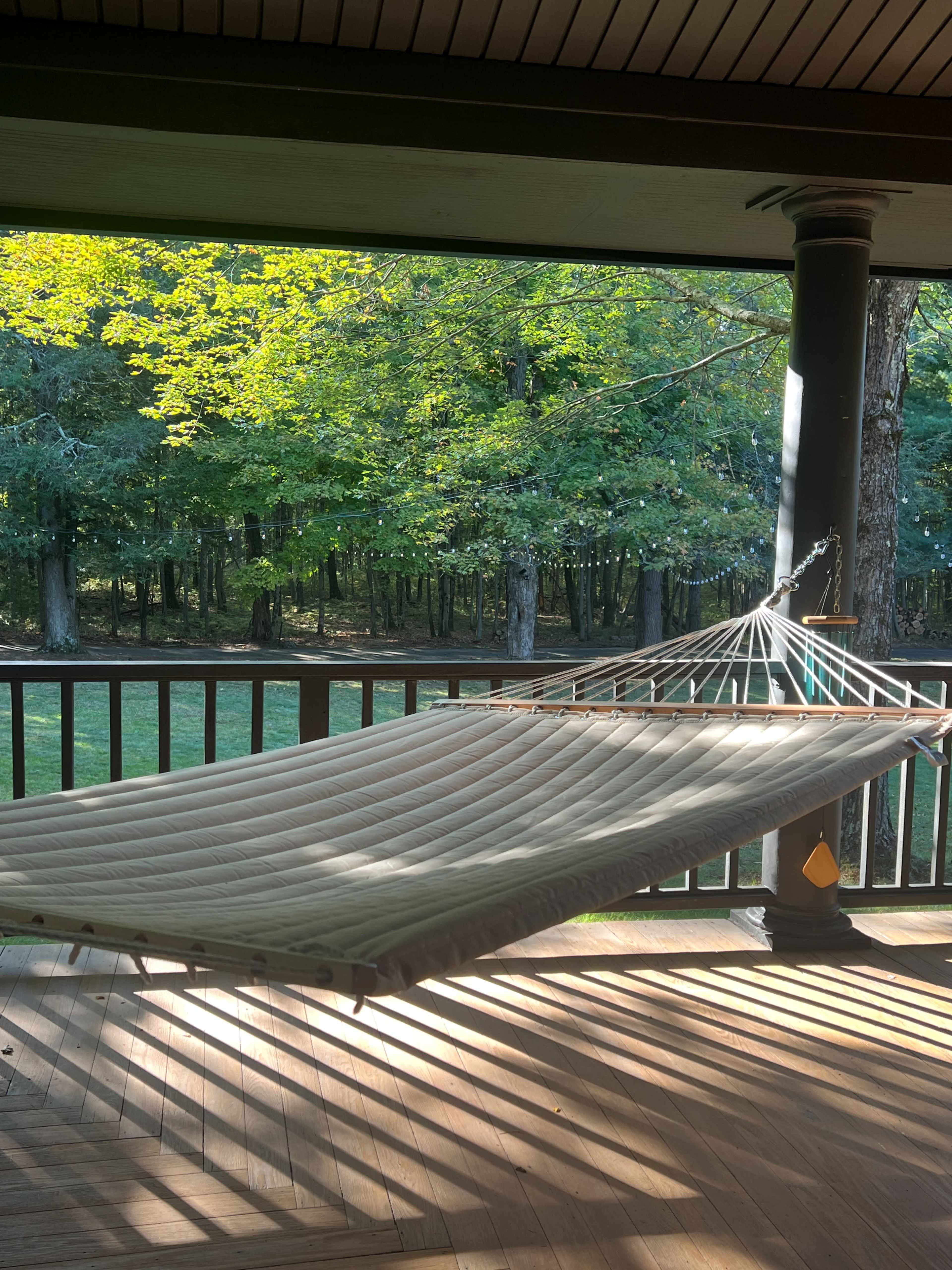 A hammock is suspended on a porch, overlooking a grassy area with trees in the background.