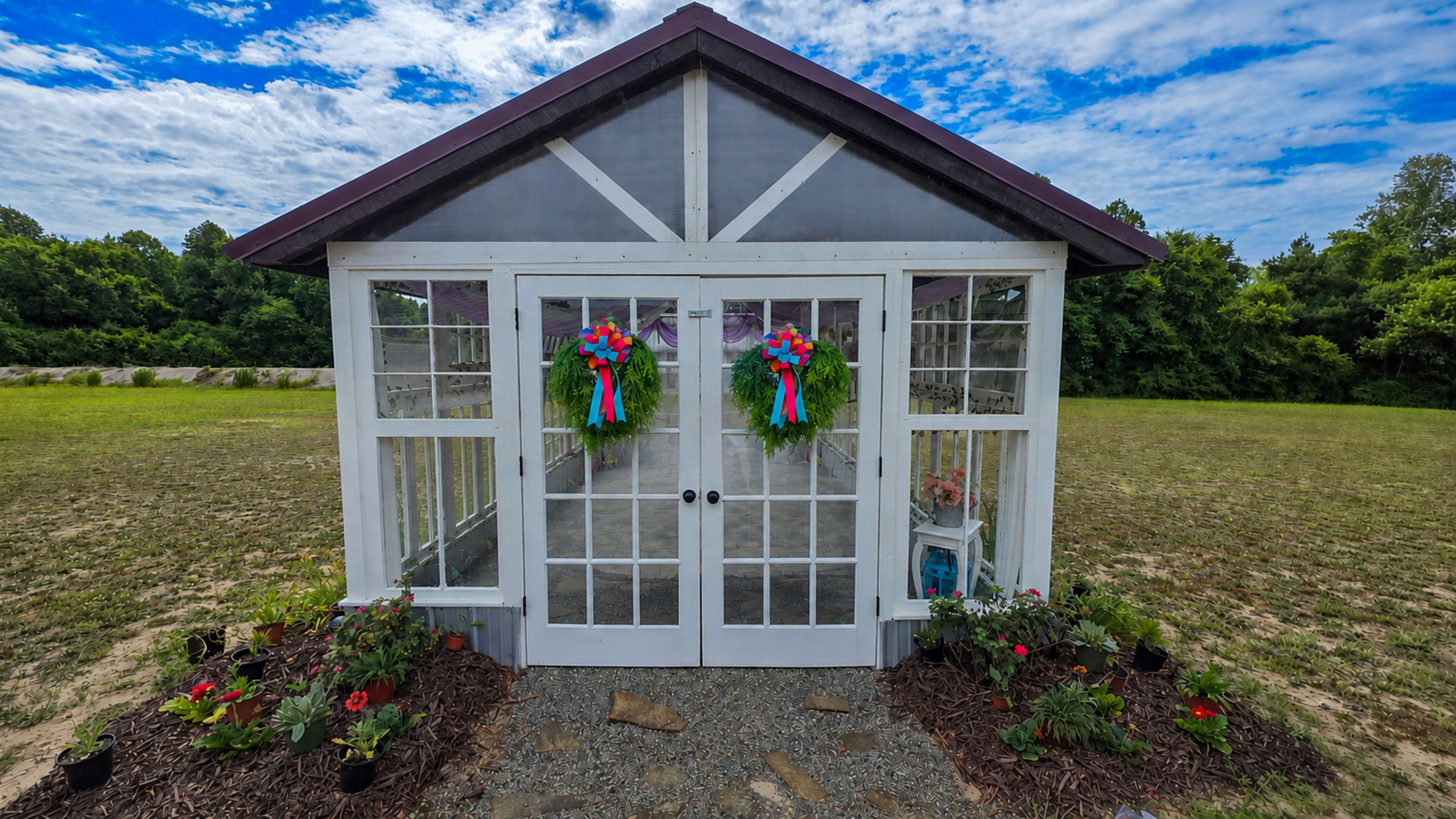 The image shows a greenhouse with double doors adorned with wreaths, surrounded by flower beds on a grassy field under a partly cloudy sky.
