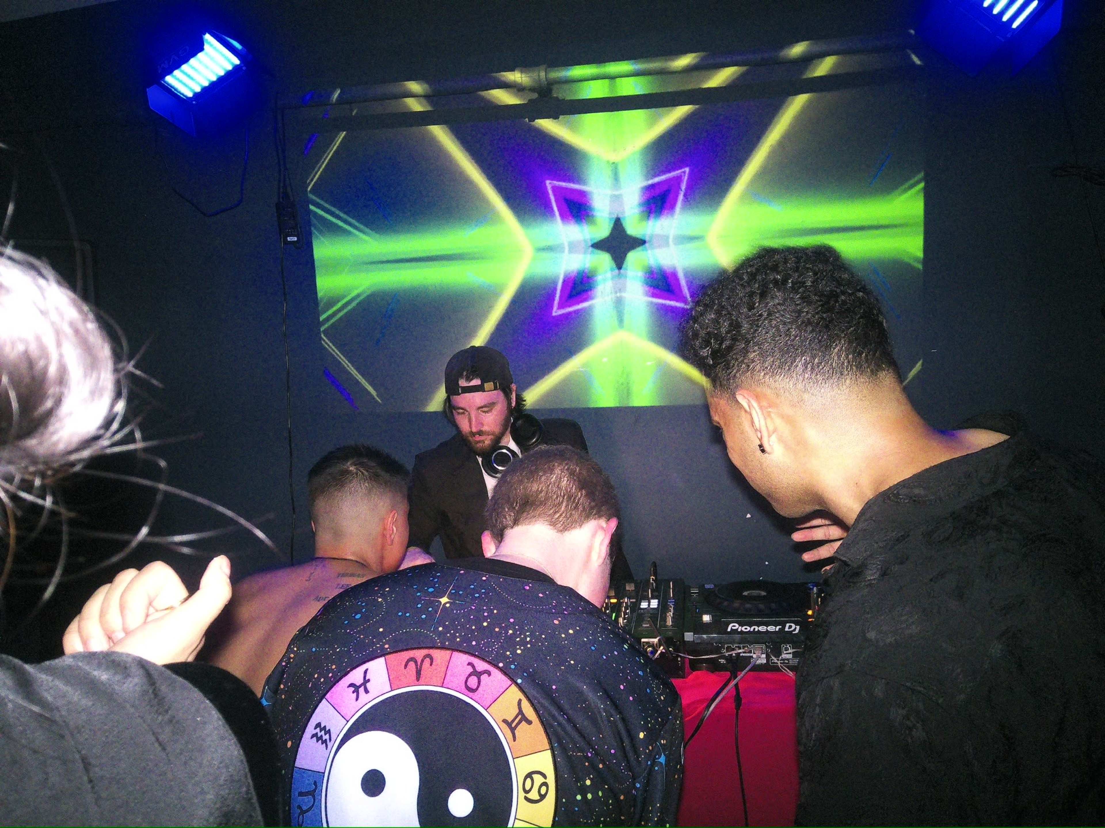 A DJ performs at a vibrant event with animated lights projected on a wall while attendees engage around the DJ booth.