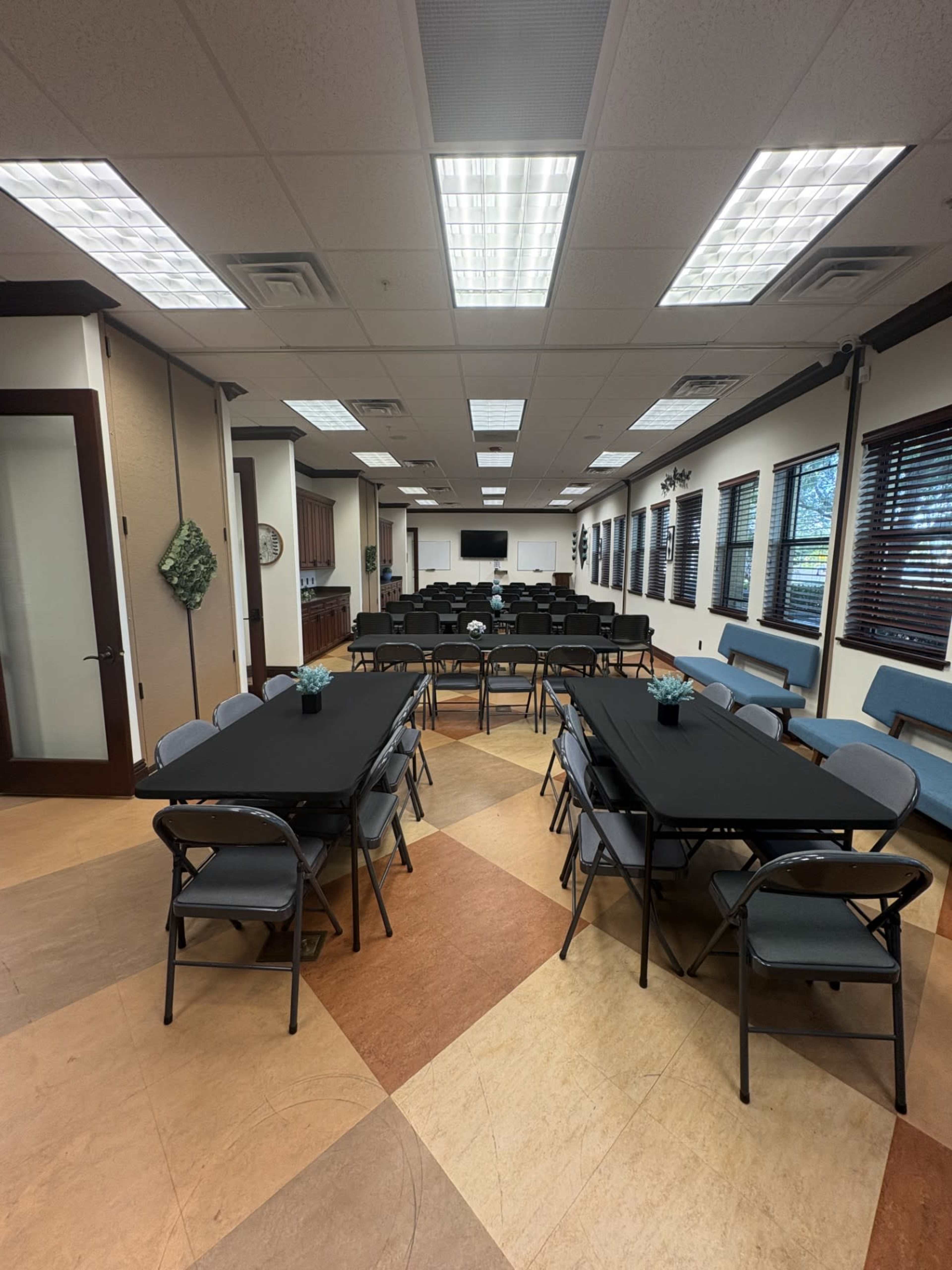 A large meeting room is set up with multiple rows of black-covered tables and folding chairs, featuring natural light from windows and a kitchenette area in the background.