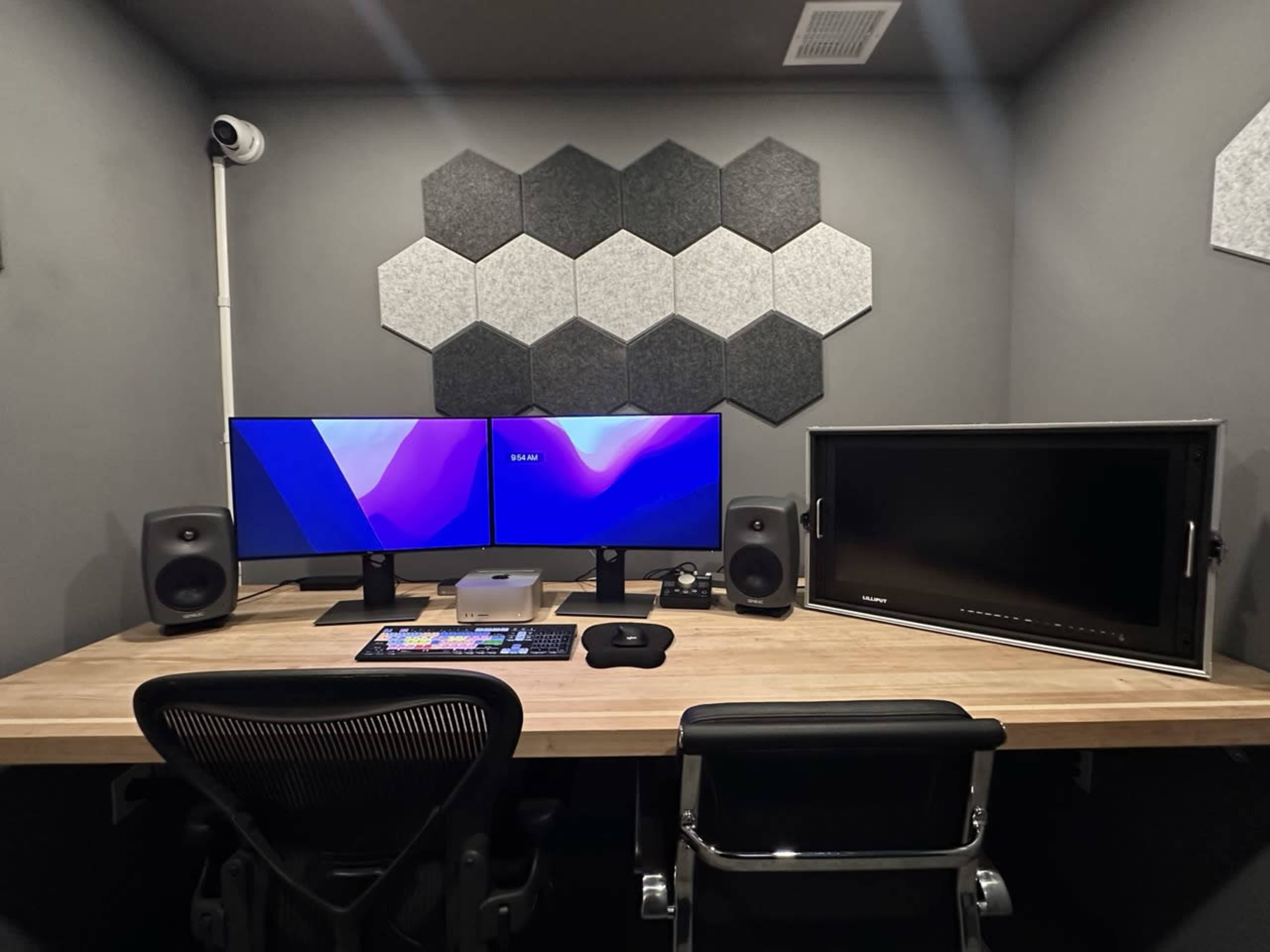 A modern workspace featuring a wooden desk with two monitors, speakers, and a keyboard, against a wall adorned with hexagonal acoustic panels.
