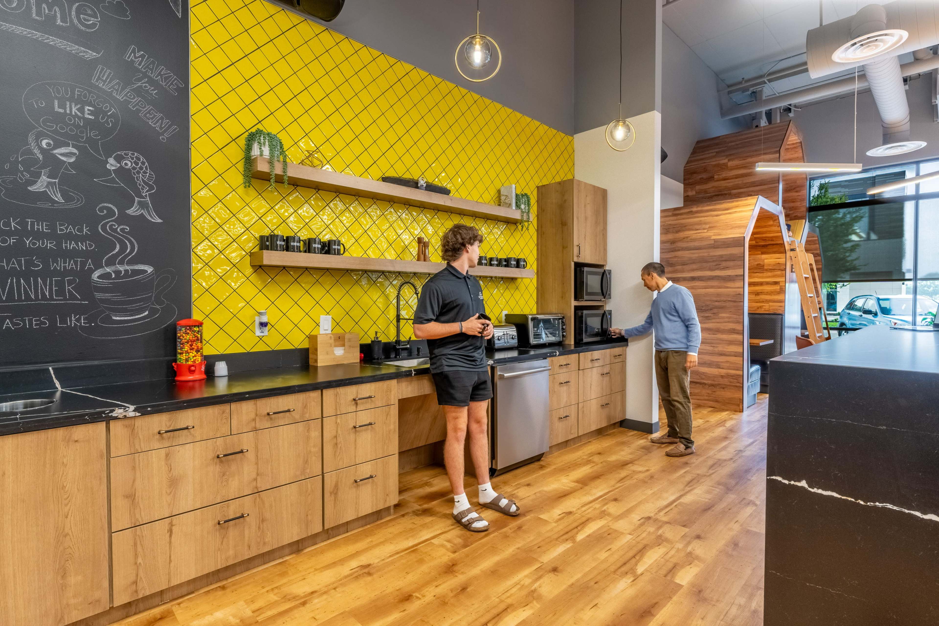 Two men stand in a modern kitchen area with wooden cabinets and a yellow tile backsplash, one looking at the microwave while the other is engaged with the coffee station.