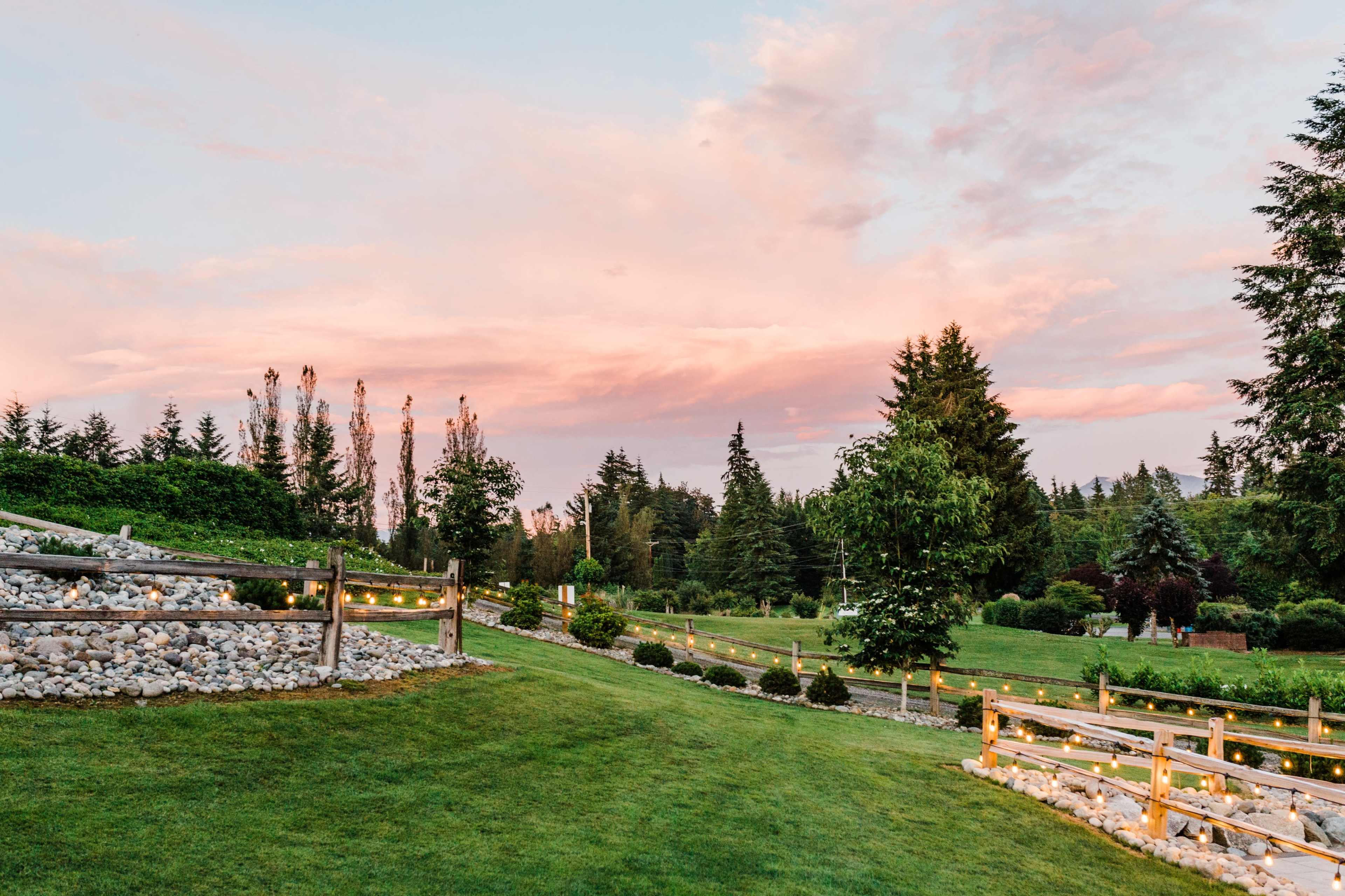 The scene depicts a landscaped area with rolling green grass, wooden fences, and trees under a pink and blue sky at sunset.