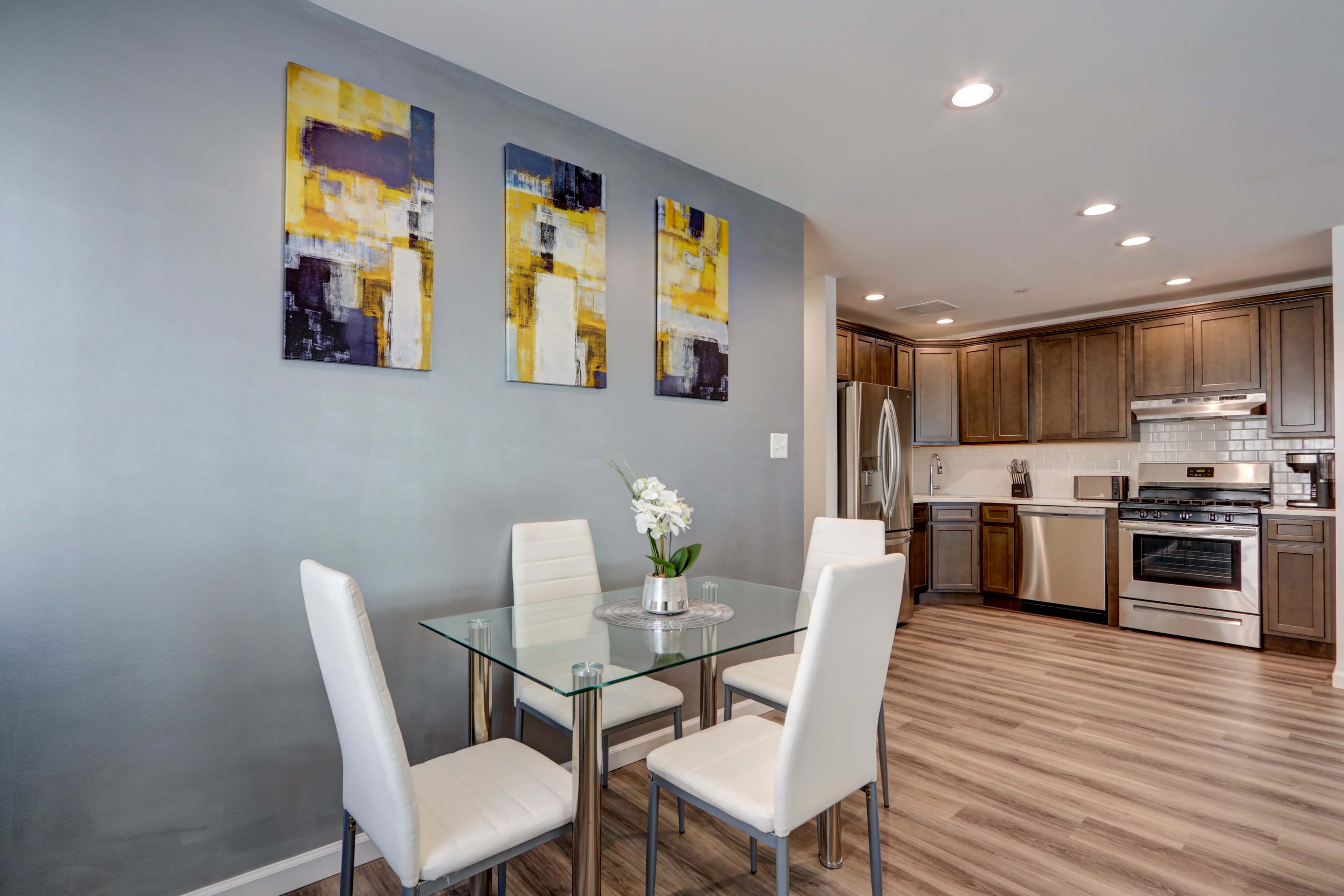 A modern kitchen and dining area featuring a glass table with four white chairs, three abstract paintings on the wall, and stainless steel appliances.