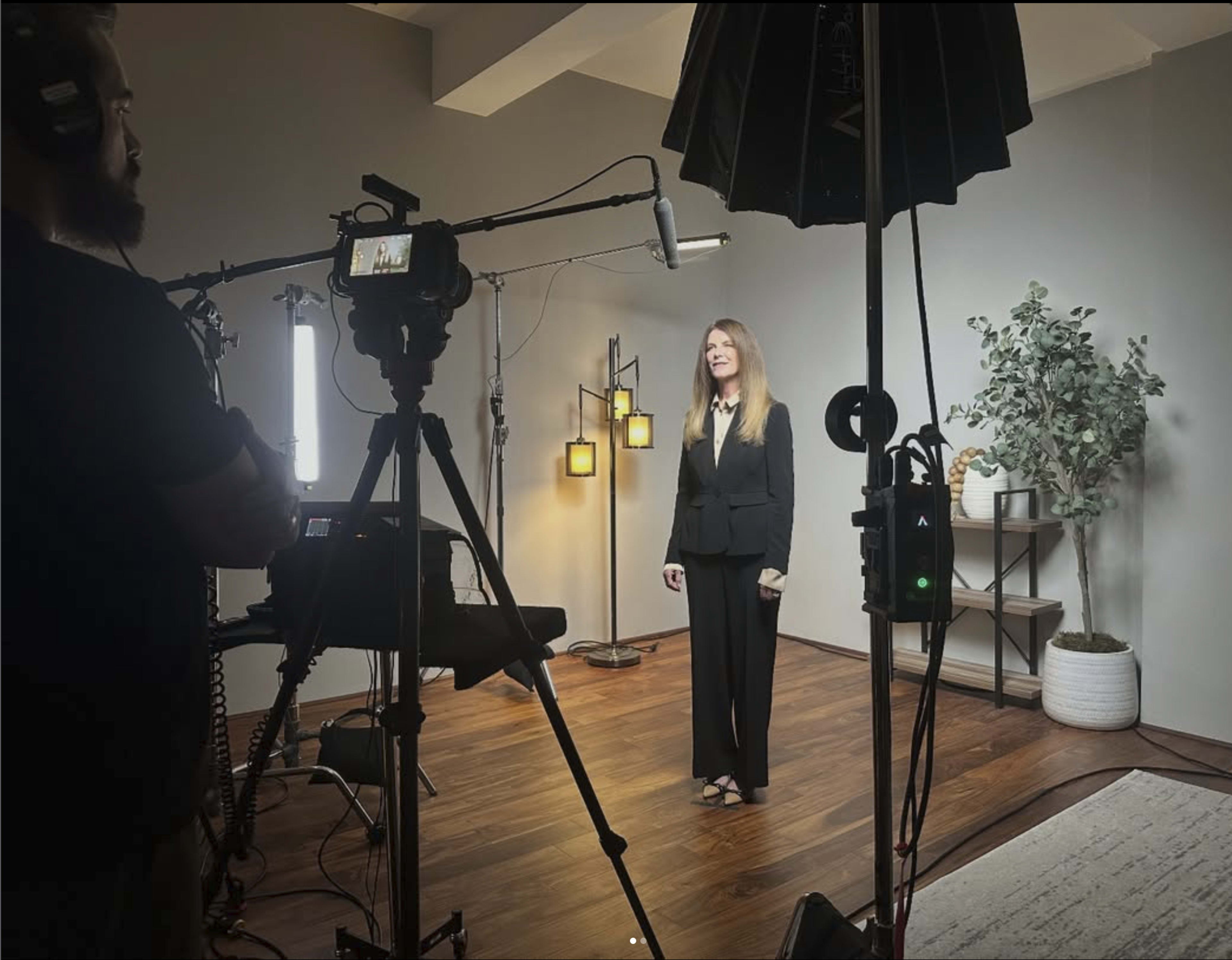 A woman in a black business suit stands in front of a camera in a well-lit studio setting with decorative lamps and plants.