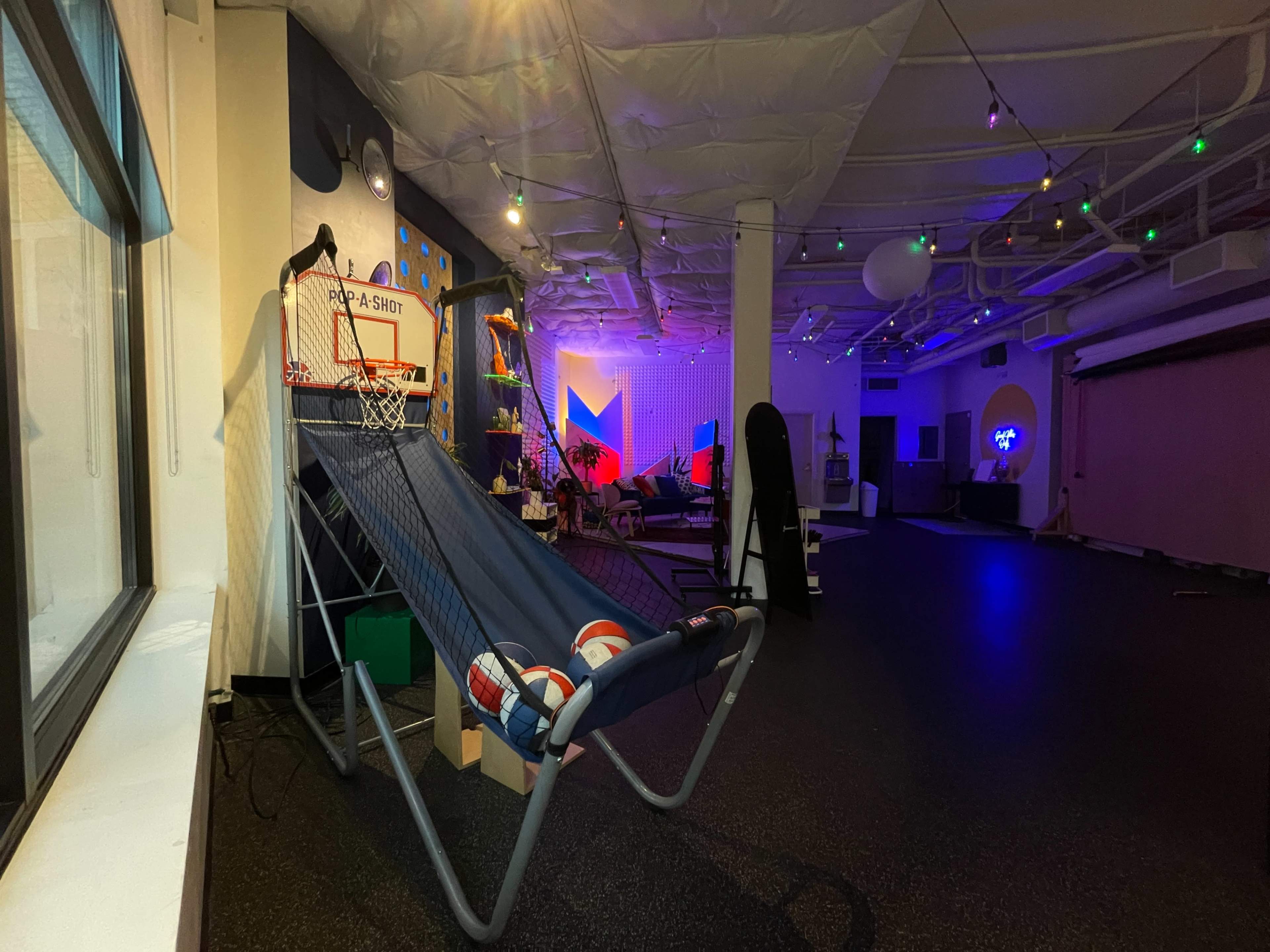 A spacious room featuring a basketball hoop set up beside a lounge chair, with colorful lighting and various recreational equipment in the background.