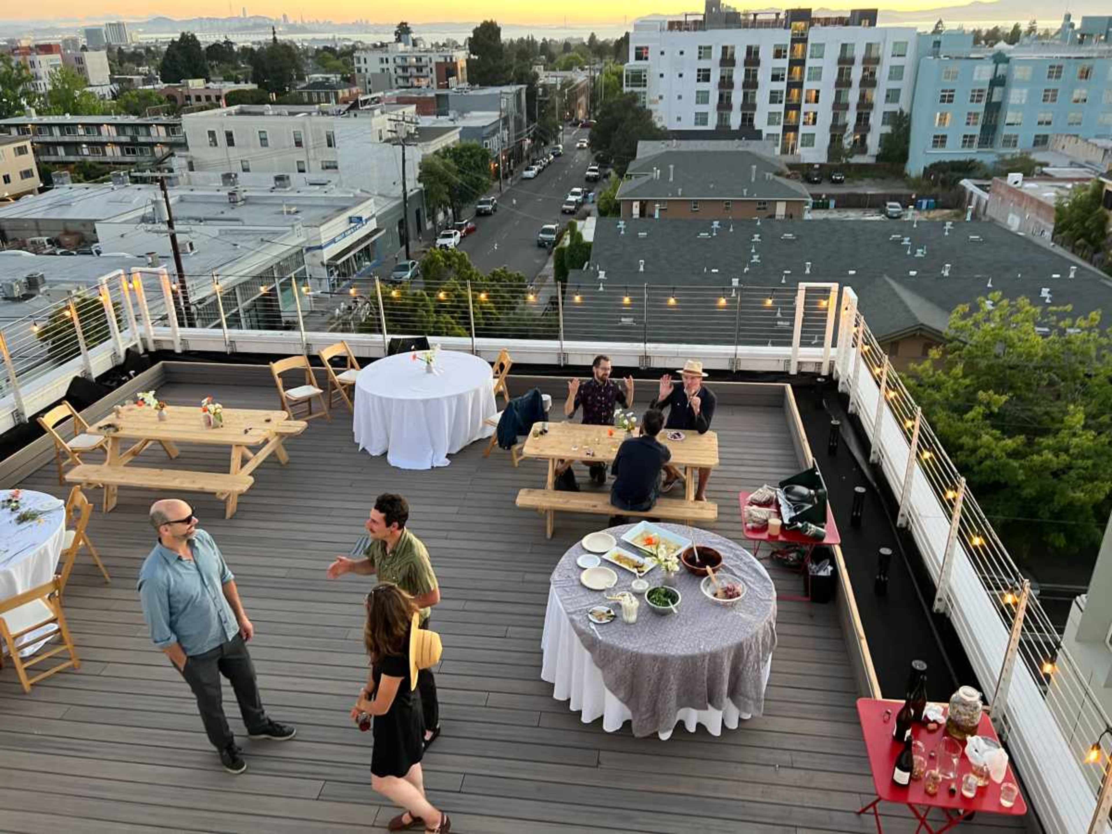 A rooftop gathering features several tables with food and drinks, while guests socialize against a backdrop of city buildings at sunset.