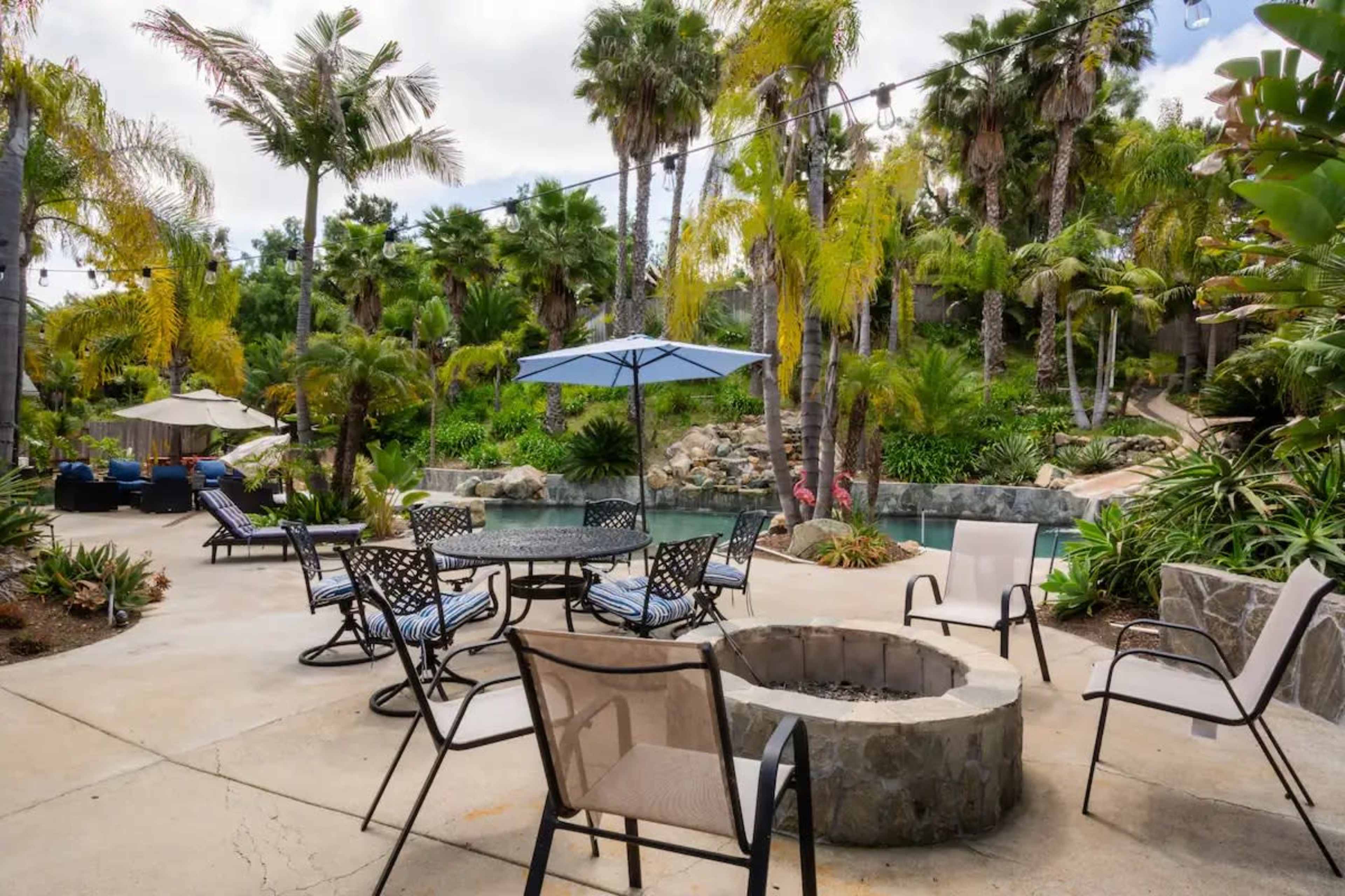 The image shows a landscaped outdoor area with a fire pit surrounded by chairs, palm trees, and a swimming pool in the background.