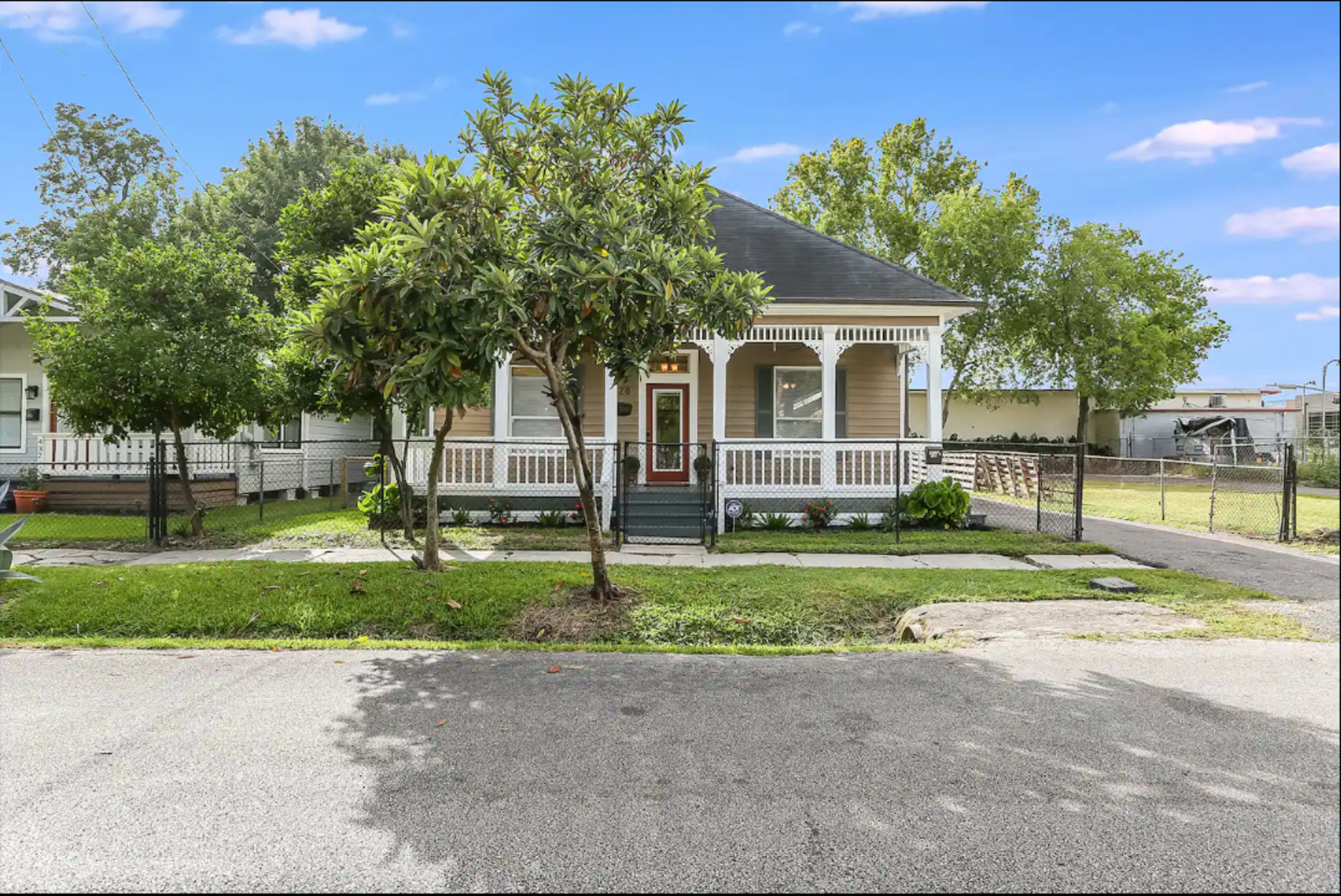 A single-story house with a porch and front yard is situated on a quiet street, surrounded by greenery and a fence.