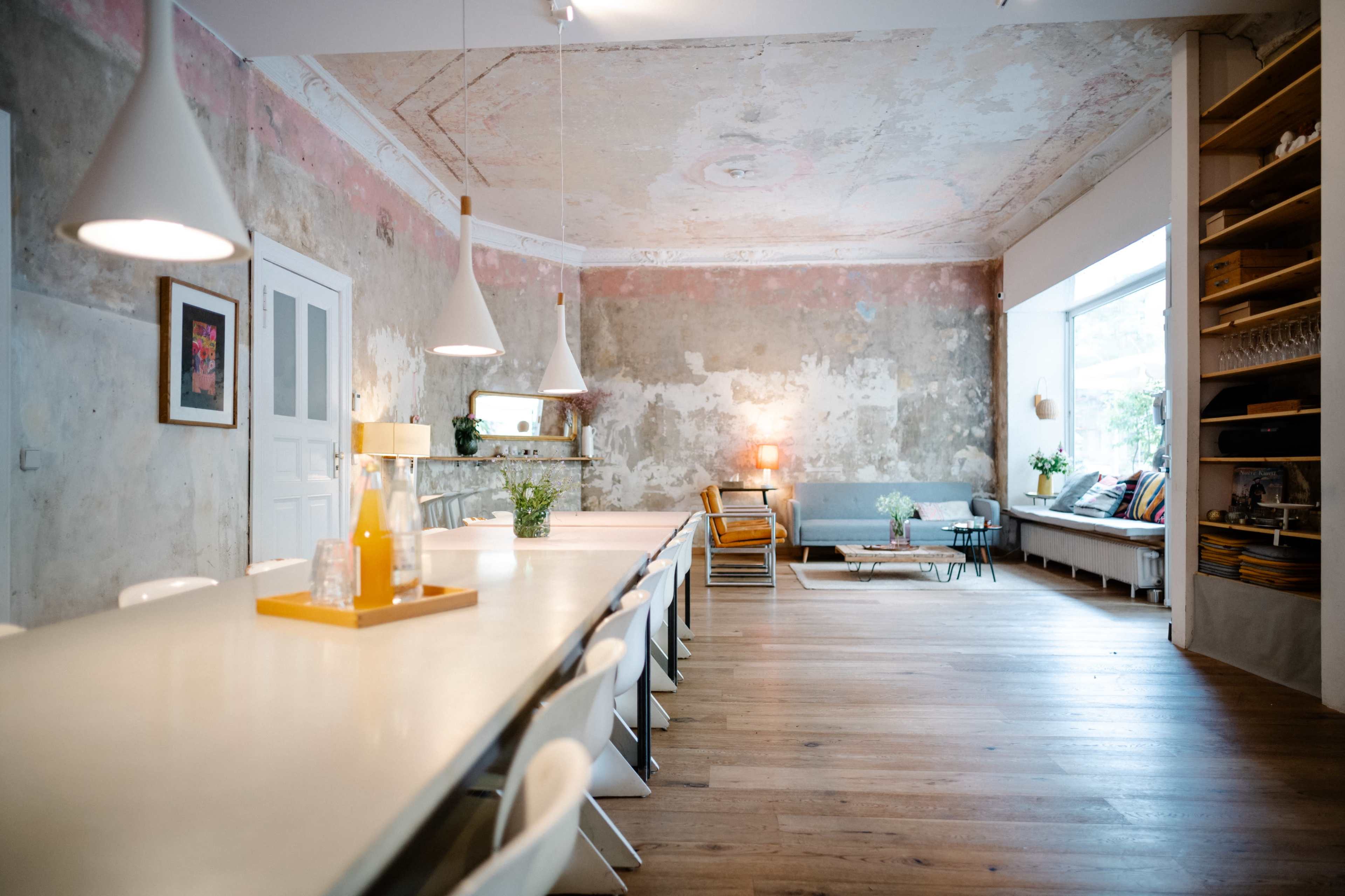 The image shows a spacious, modern dining area with a long white table, white chairs, and exposed brick walls, complemented by natural light from a large window.