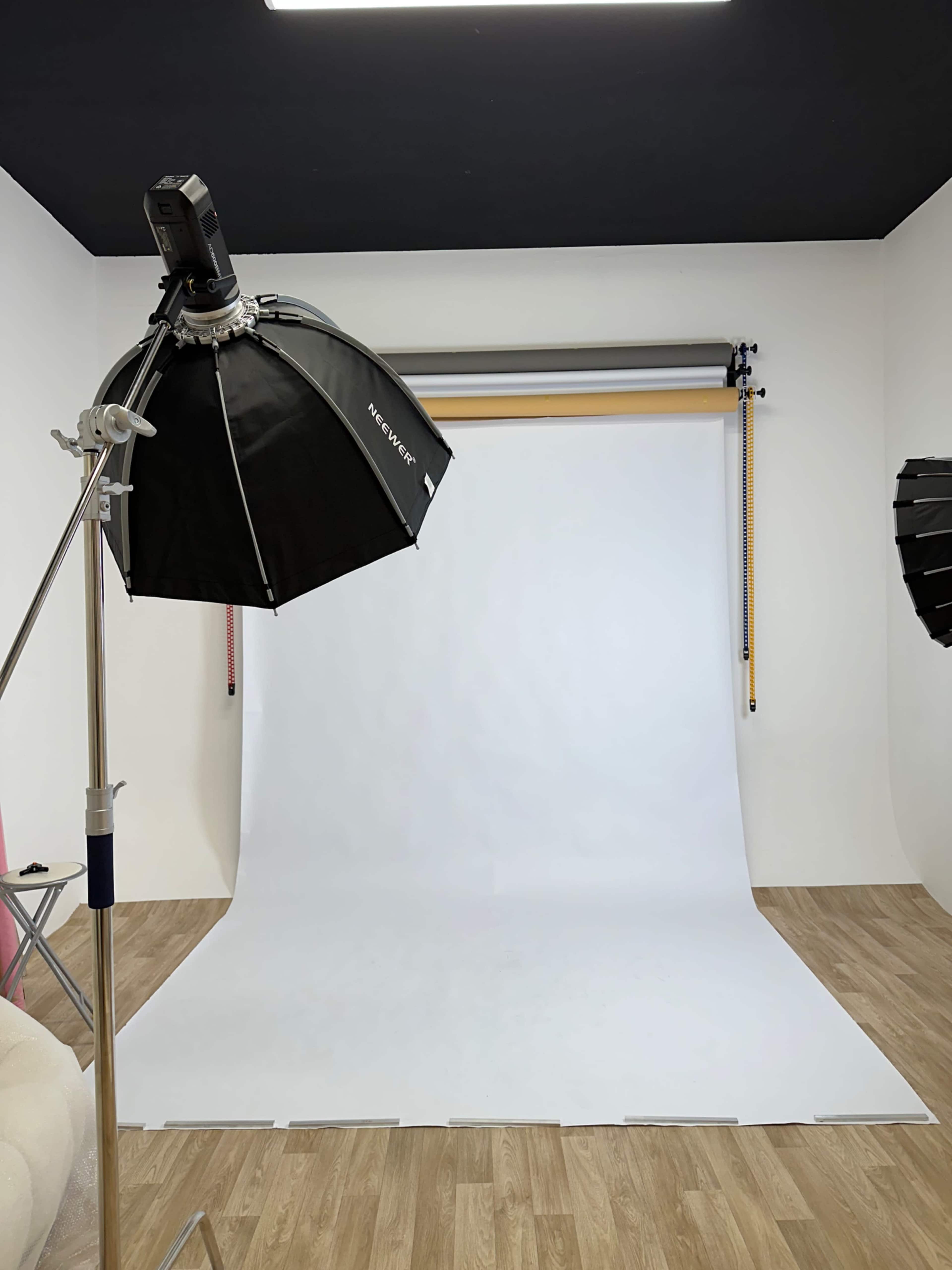 The image shows a photography studio with a white backdrop and lighting equipment set up in a well-lit room.