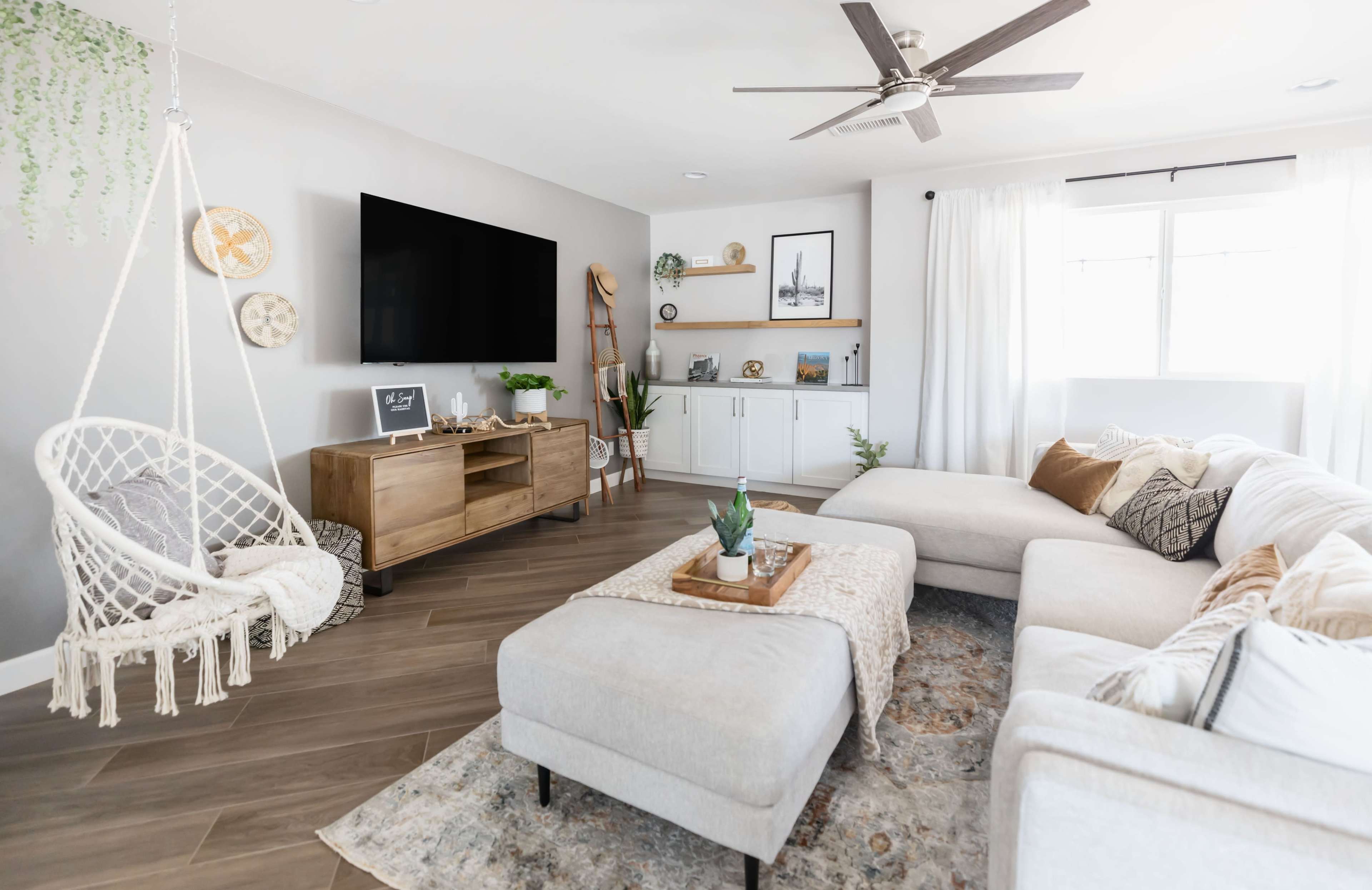The image shows a modern living room with a white sectional sofa, a hanging chair, a wooden TV stand, and a coffee table decorated with a plant and a tray.