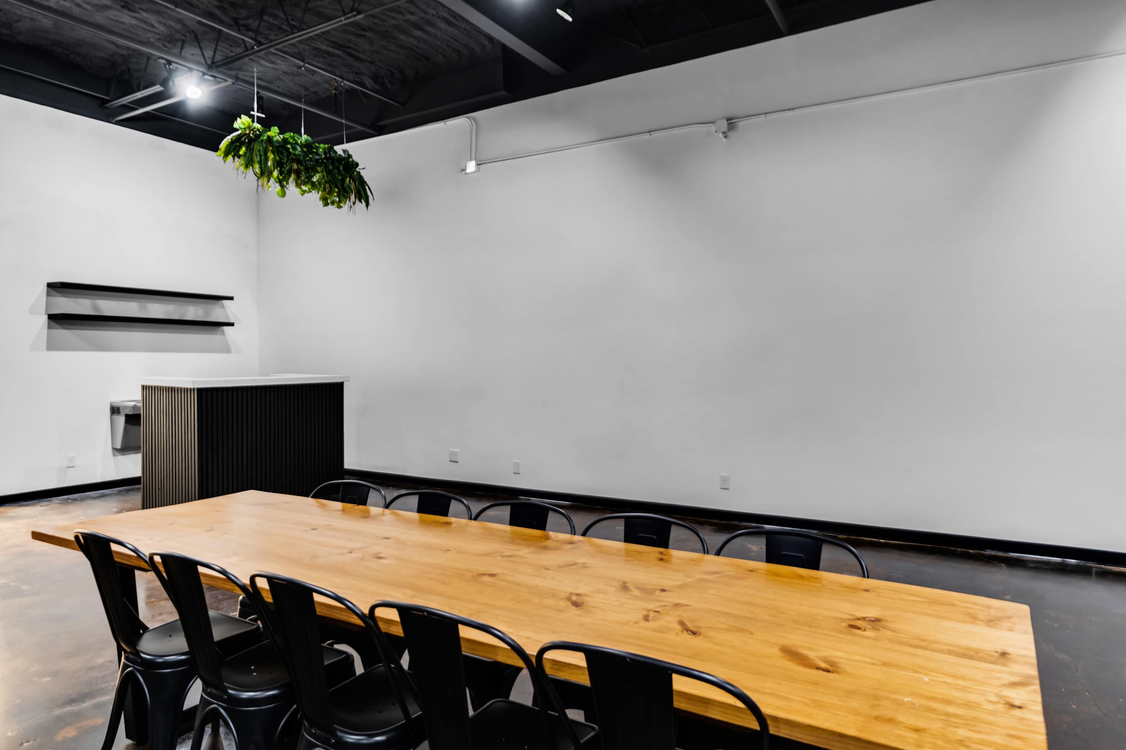 A long wooden table with black chairs is positioned in a minimalist room featuring a hanging plant and a dark storage unit against the wall.