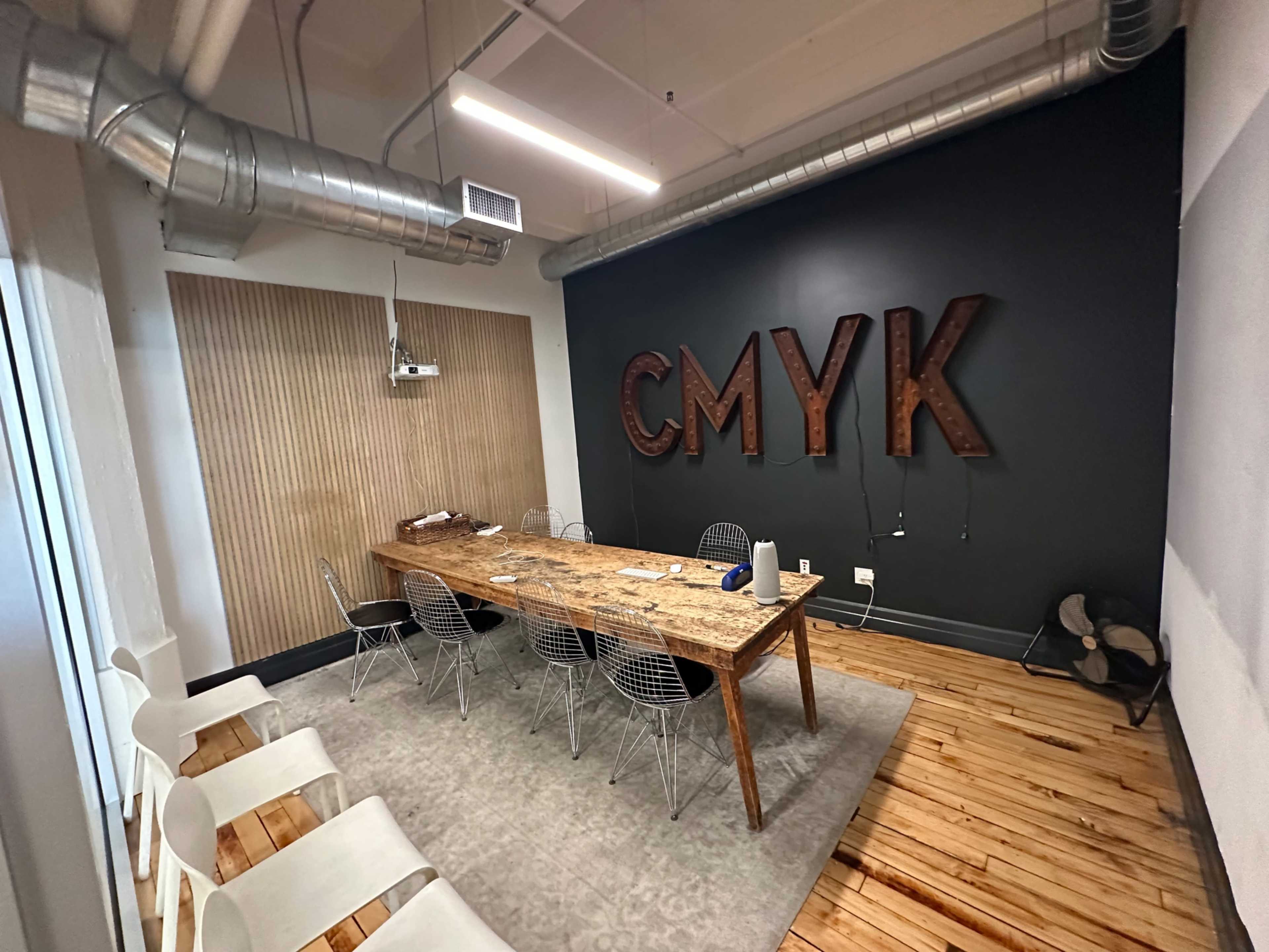 The image shows a conference room featuring a wooden table with six chairs, a large black wall displaying the letters "CMYK," and a light-colored rug on the floor.