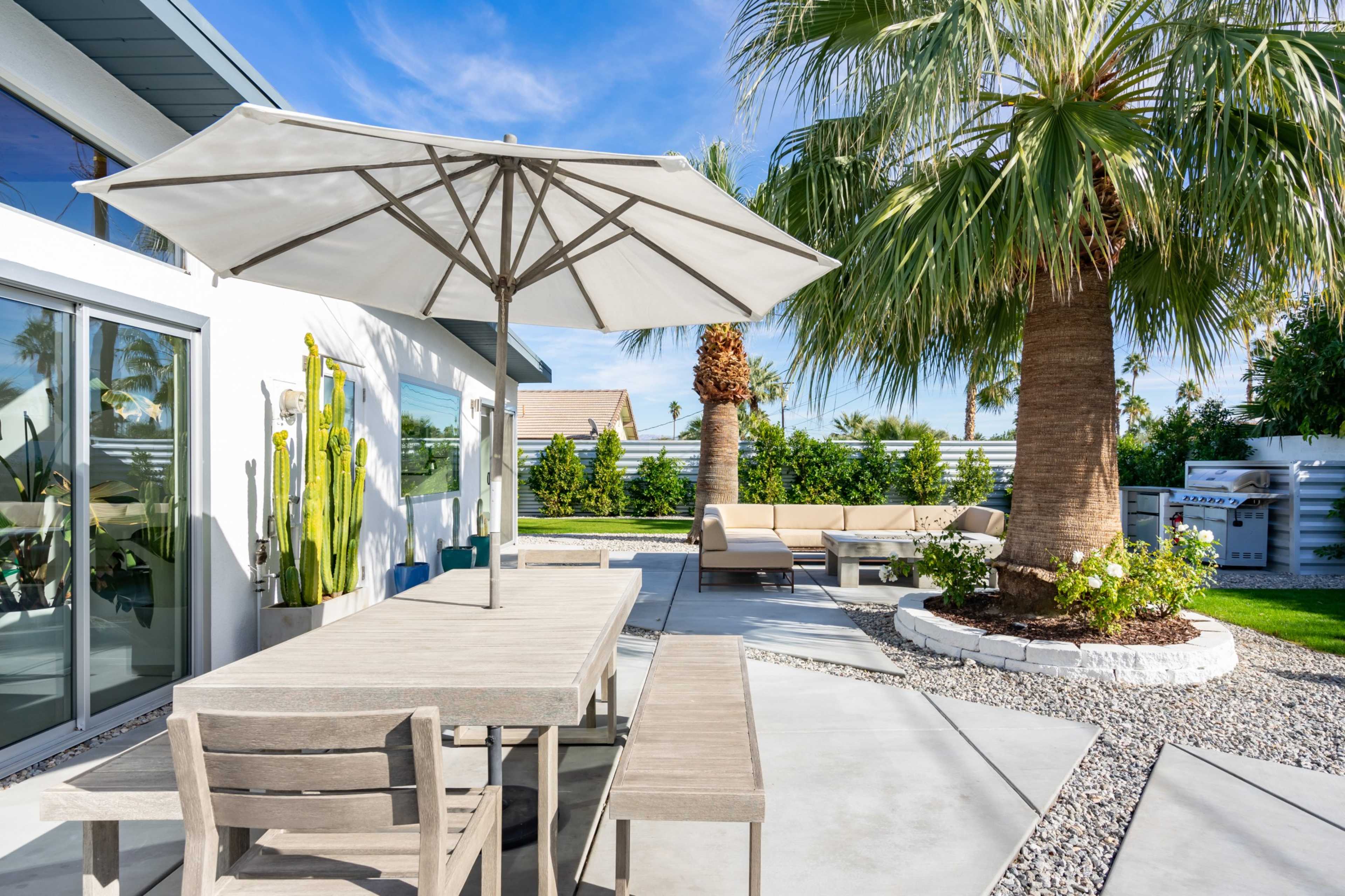 A modern outdoor patio features a dining table with chairs, a large umbrella, and a seating area surrounded by palm trees and landscaping.