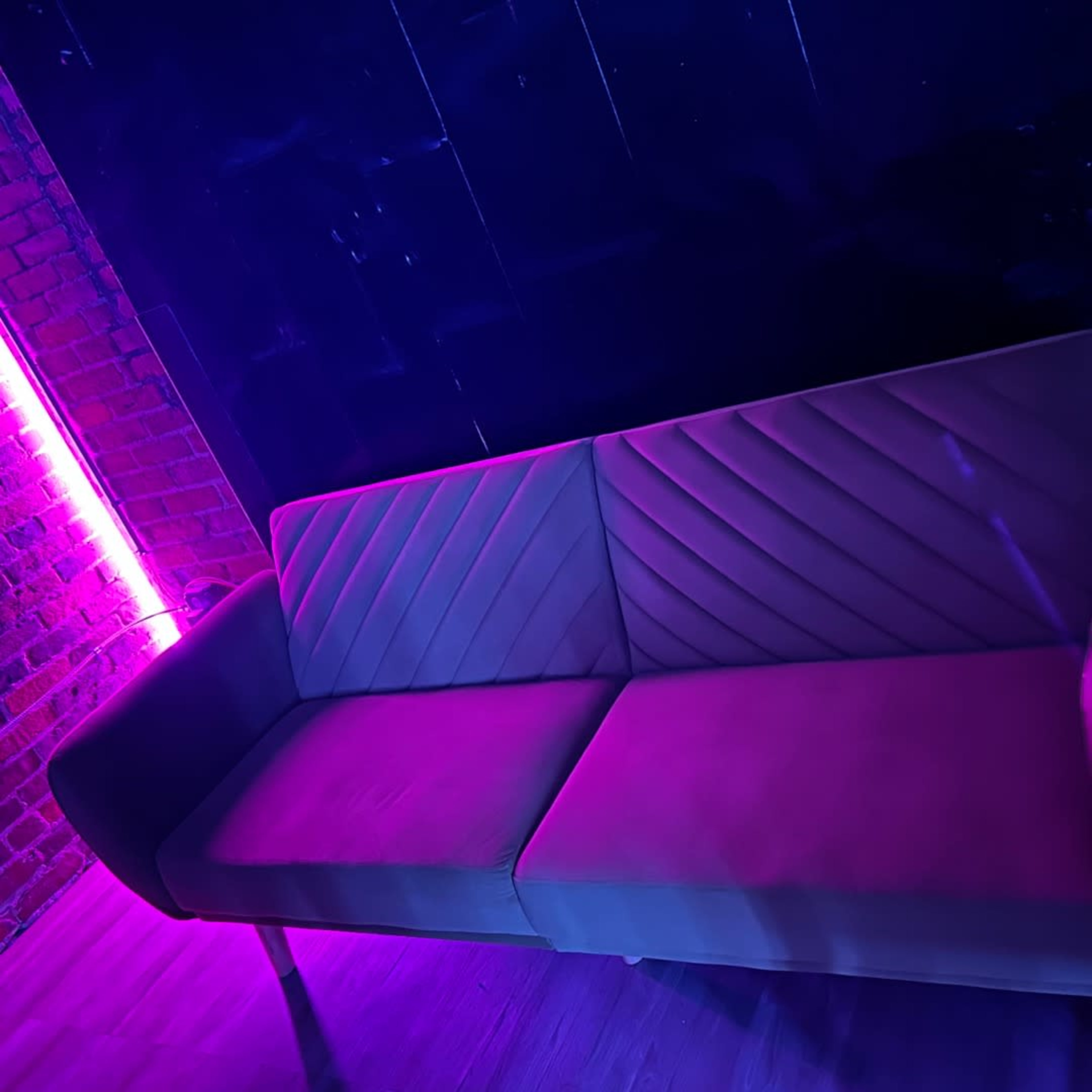 A modern, upholstered couch with a geometric pattern is positioned against a brick wall illuminated by purple neon lights.
