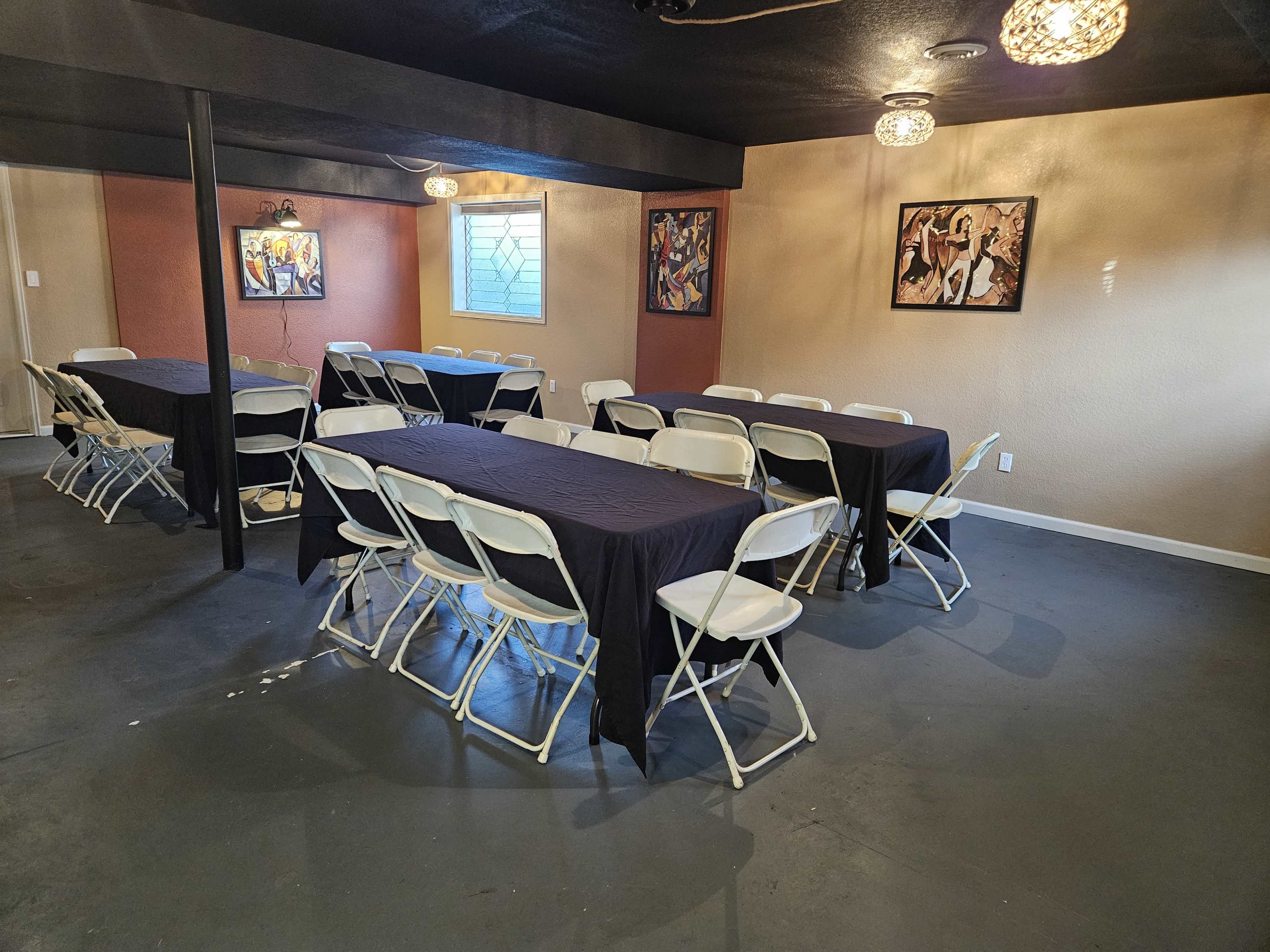 A basement room with several long tables covered in black tablecloths and white folding chairs arranged around them, alongside colorful artwork on the walls.