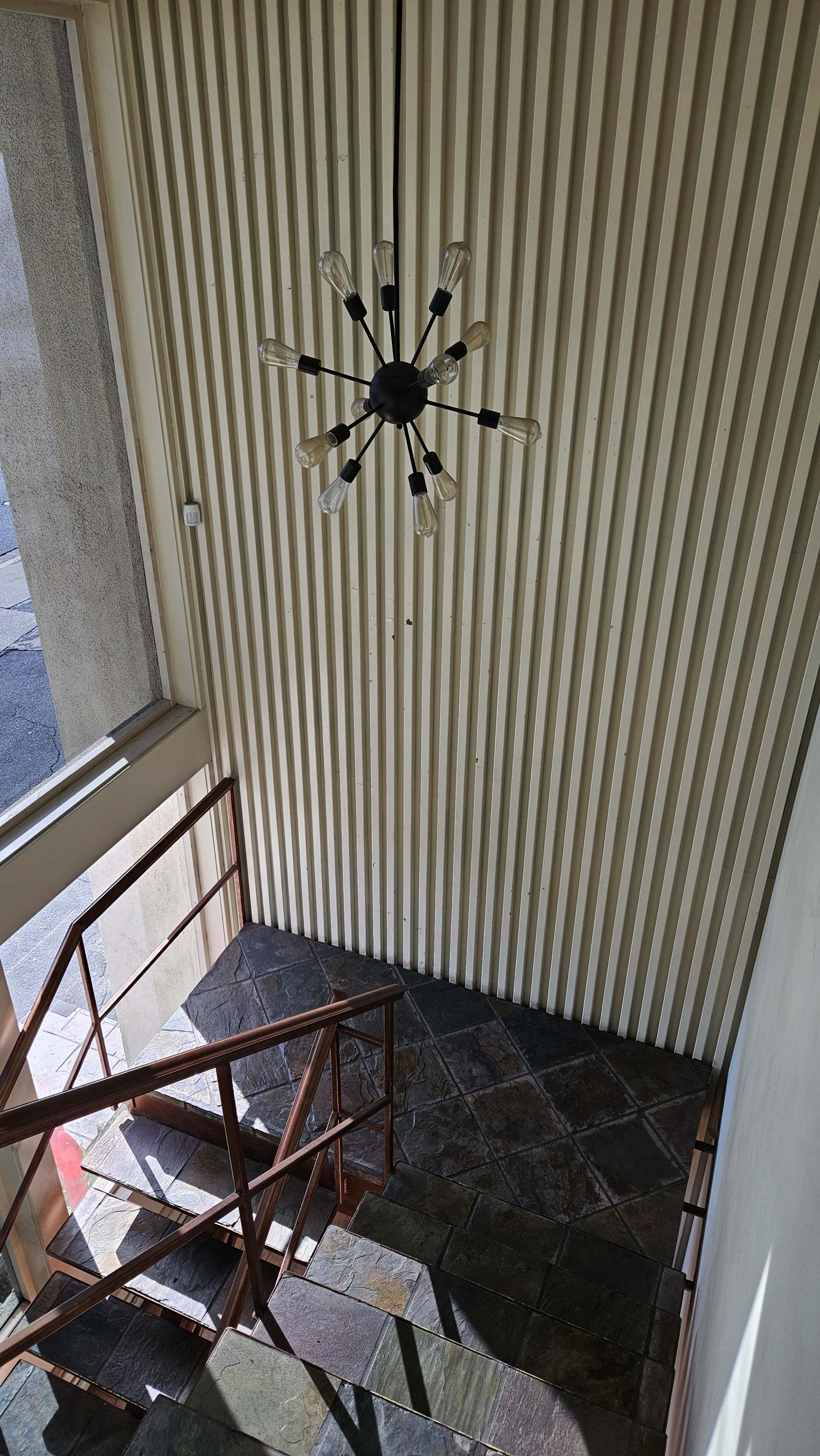 A narrow staircase descending in a lit area with vertical lined walls and a modern chandelier overhead.