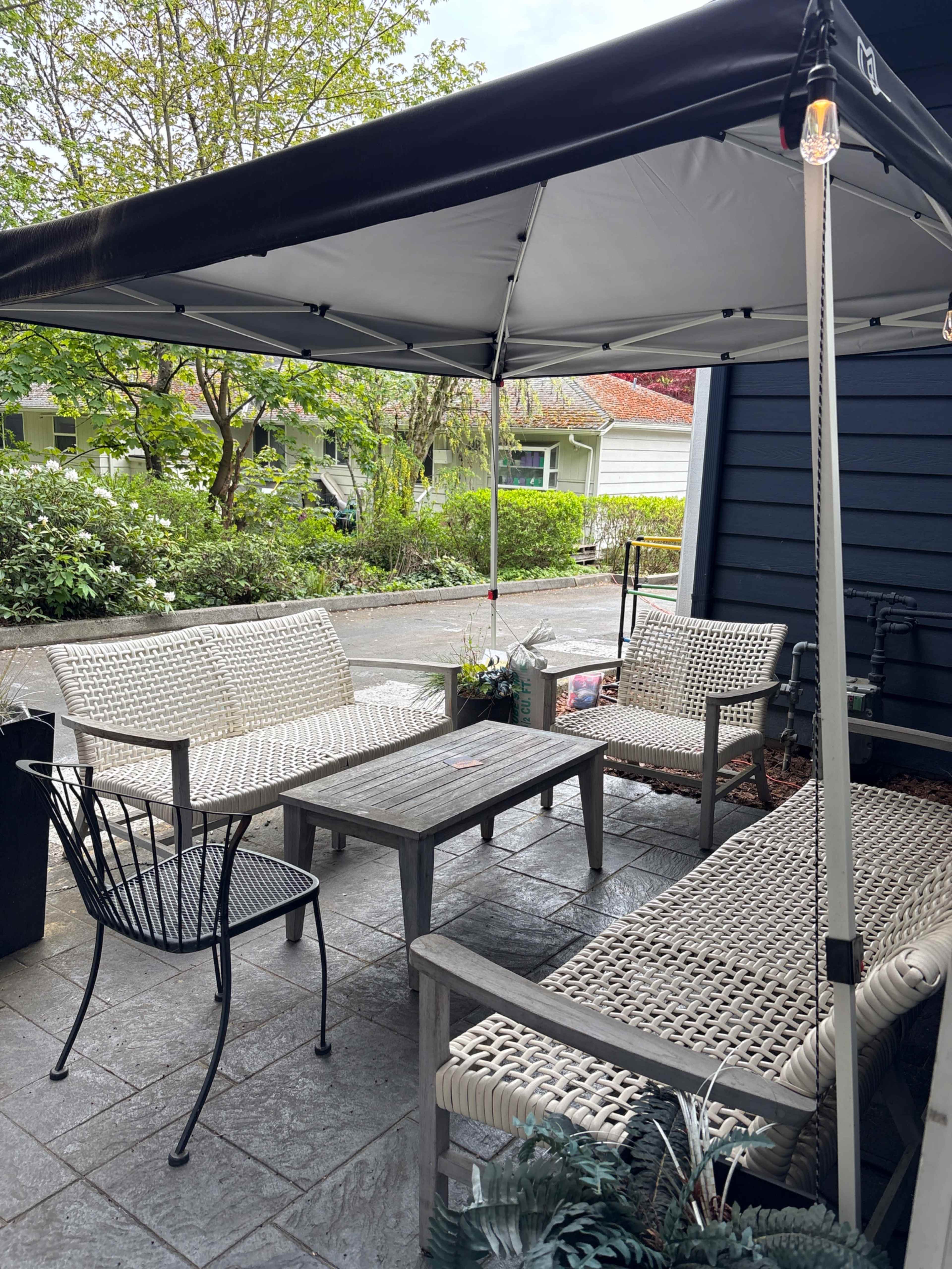 A covered outdoor seating area features two wicker chairs and a table on a stone patio surrounded by greenery.