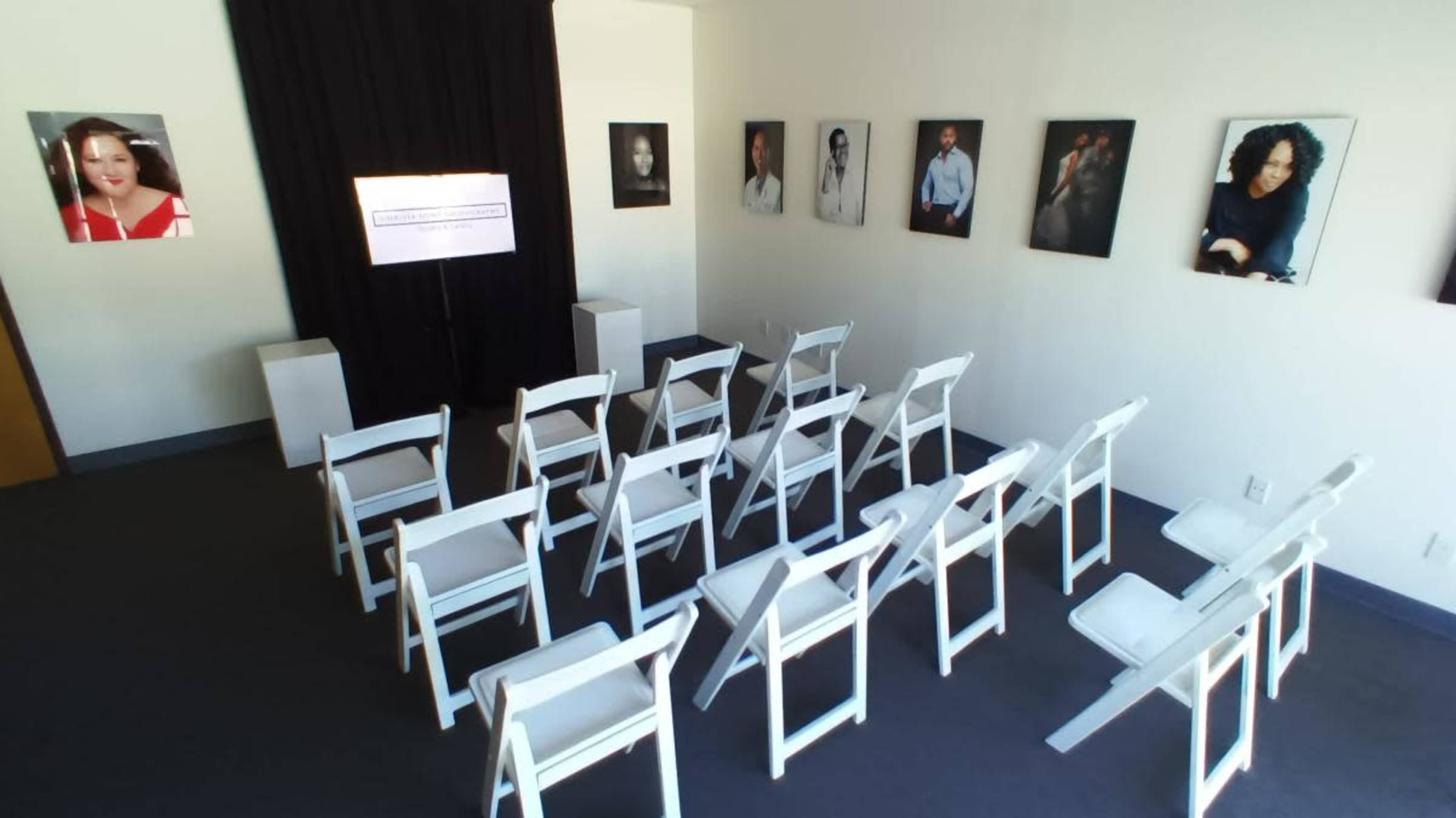 The image shows a room set up for an event with empty white chairs arranged in rows facing a screen, and black curtains in the background, adorned with framed portraits on the walls.
