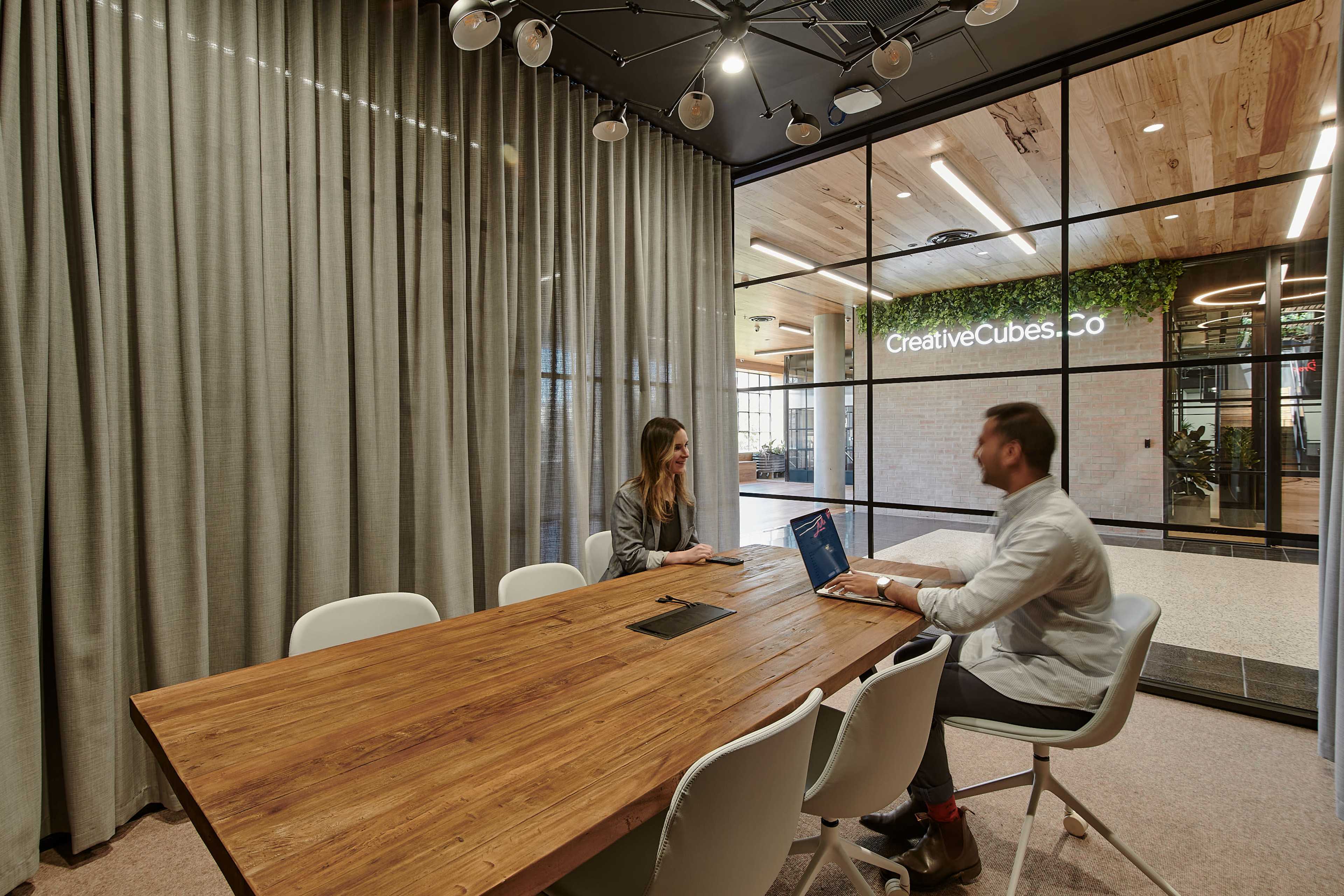 Two people are seated at a large wooden conference table in a modern meeting room with glass walls and a sign for CreativeCubes.co in the background.