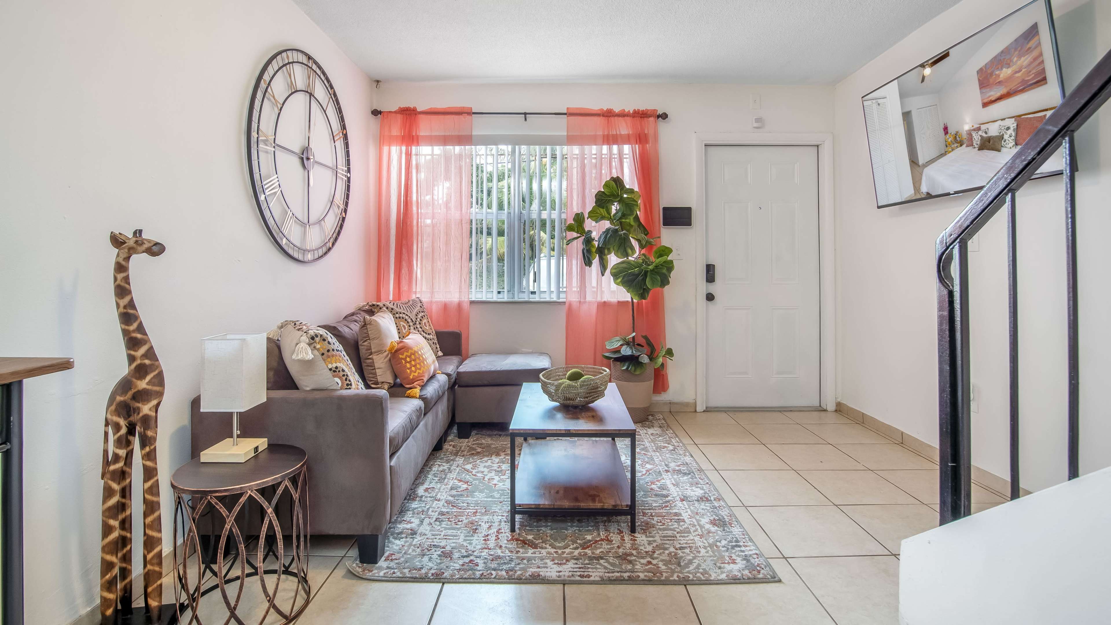 A living room with a gray sofa, a coffee table, a decorative plant, and a large wall clock, complemented by a window with orange curtains.