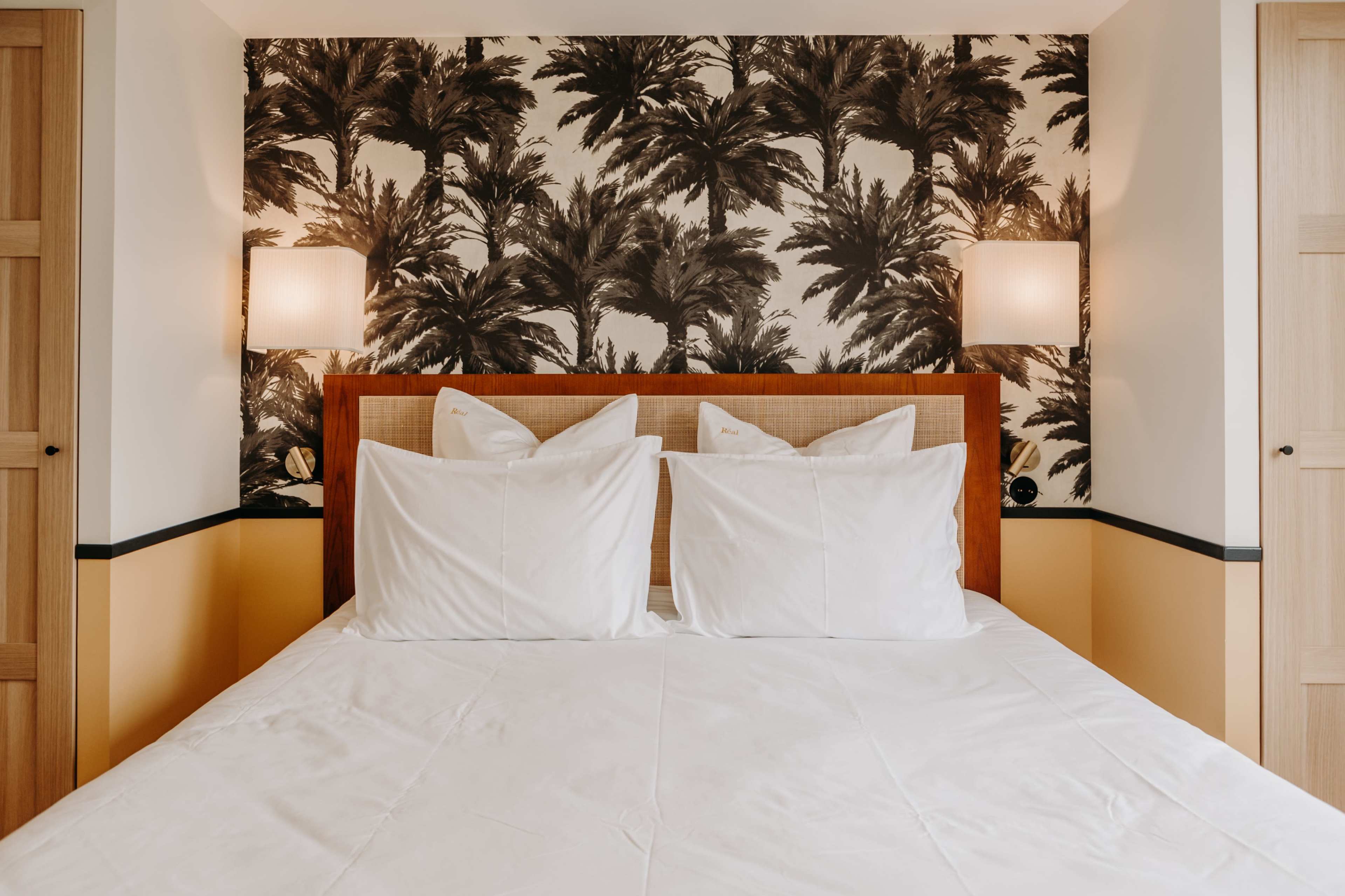 A bedroom with a bed featuring white pillows against a backdrop of palm-patterned wallpaper.