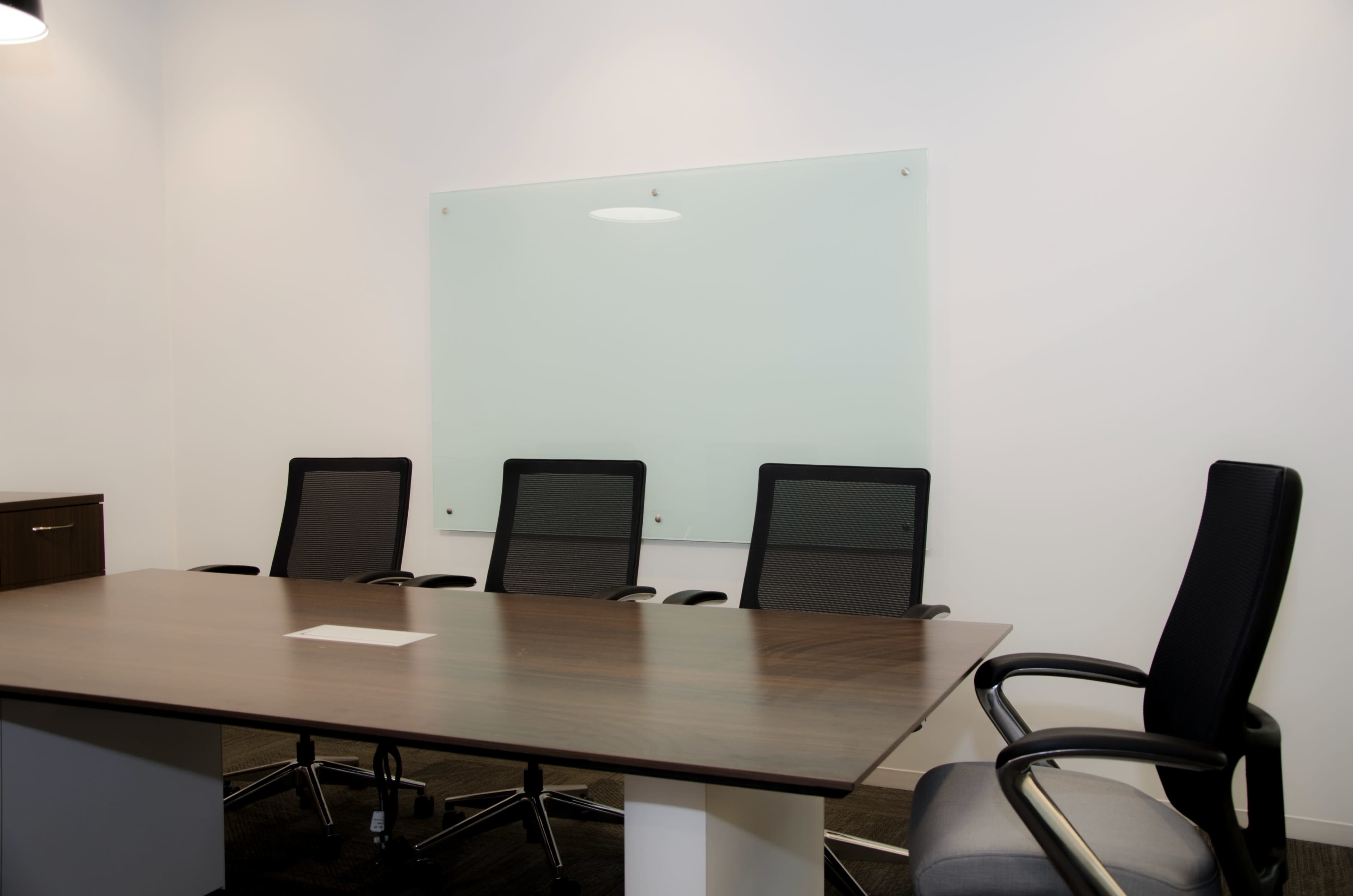A modern conference room with a wooden table and four black mesh chairs facing a large whiteboard on the wall.