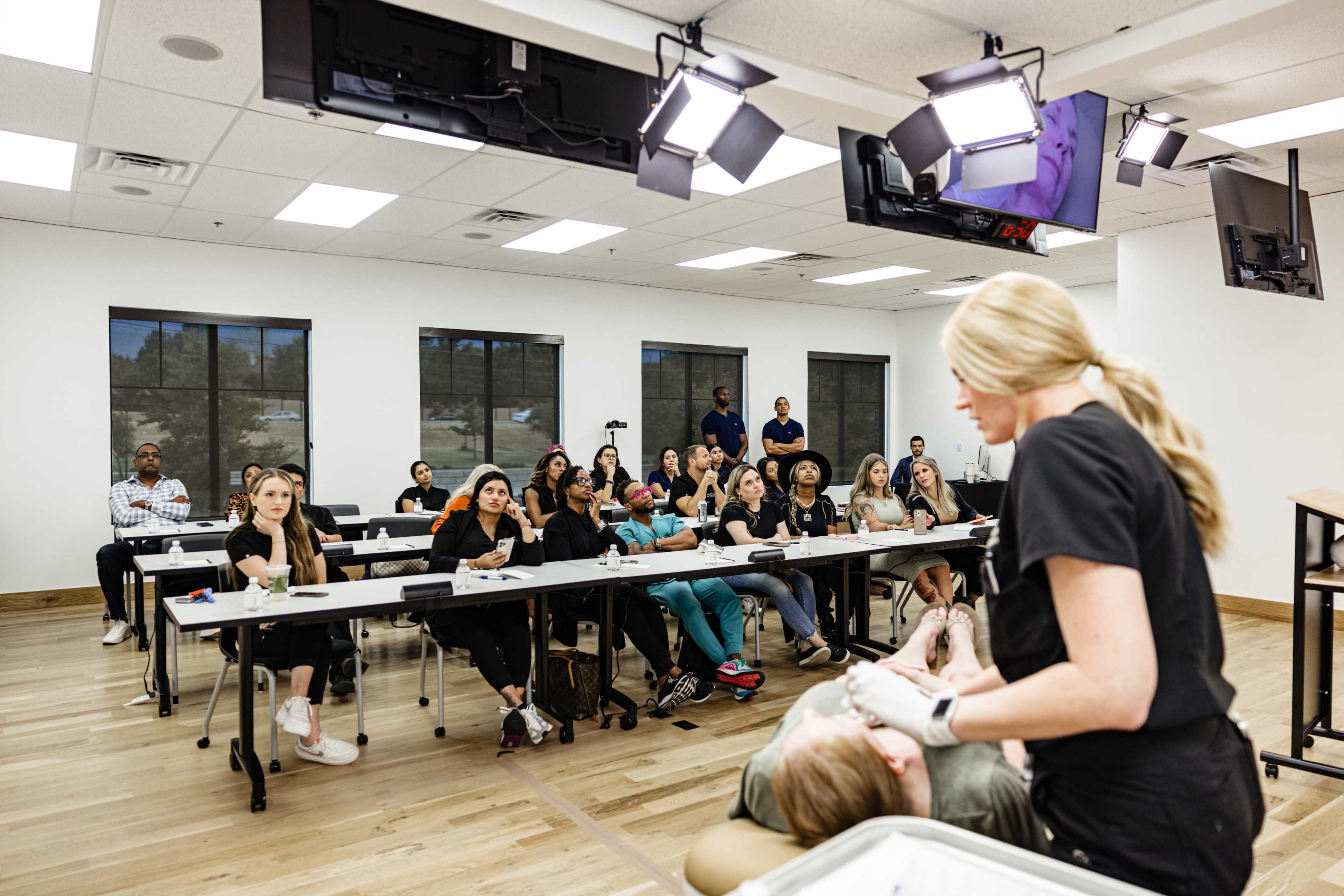 A medical or educational seminar is taking place in a classroom, where a speaker demonstrates a technique on a seated participant while an audience observes.