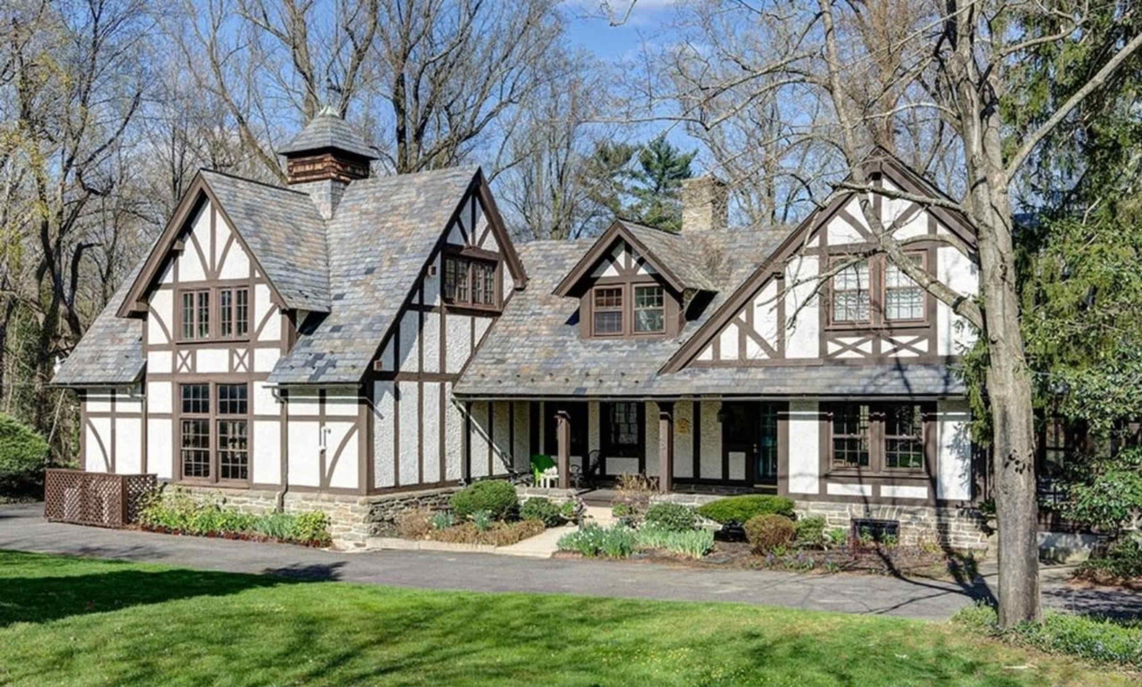 The image features a large, two-story house with a steeply pitched roof, exposed wooden beams, and a stone foundation, surrounded by greenery.