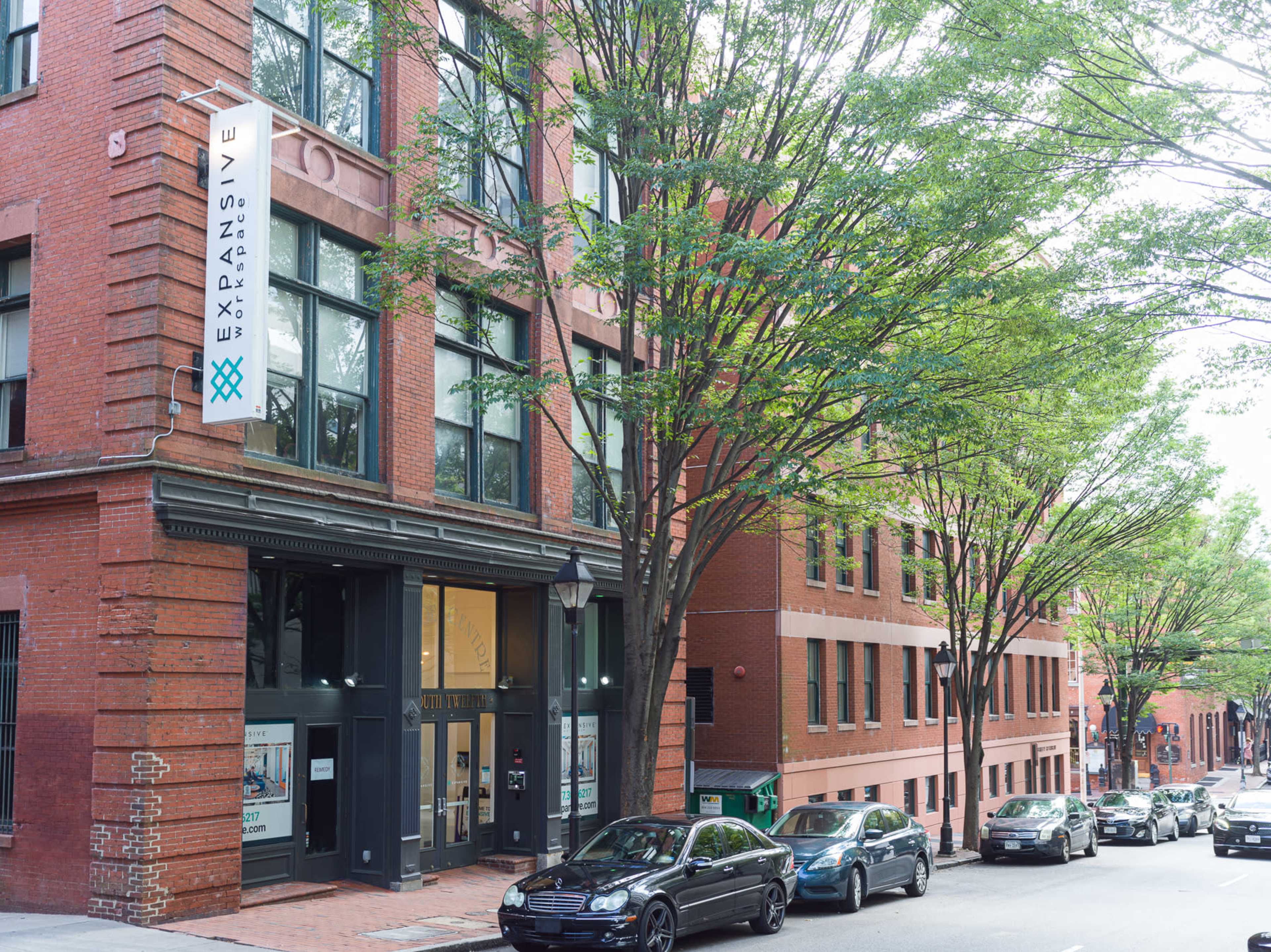 A brick building with a large sign reading "EXPANSIVE" stands next to lined trees and parked cars on a city street.