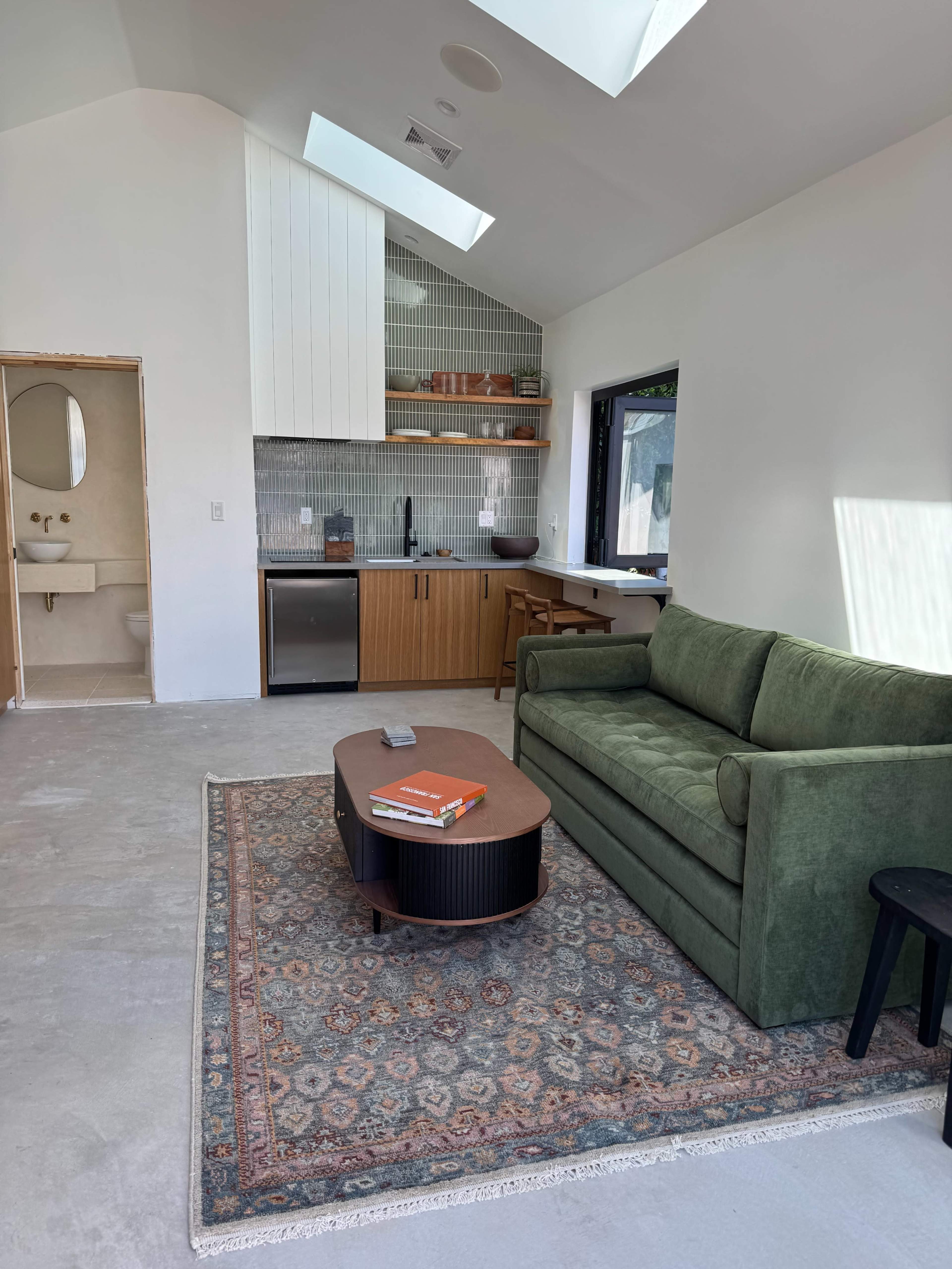 A modern living area features a green sofa, a coffee table, and a kitchenette with wooden shelves and a visible sink.
