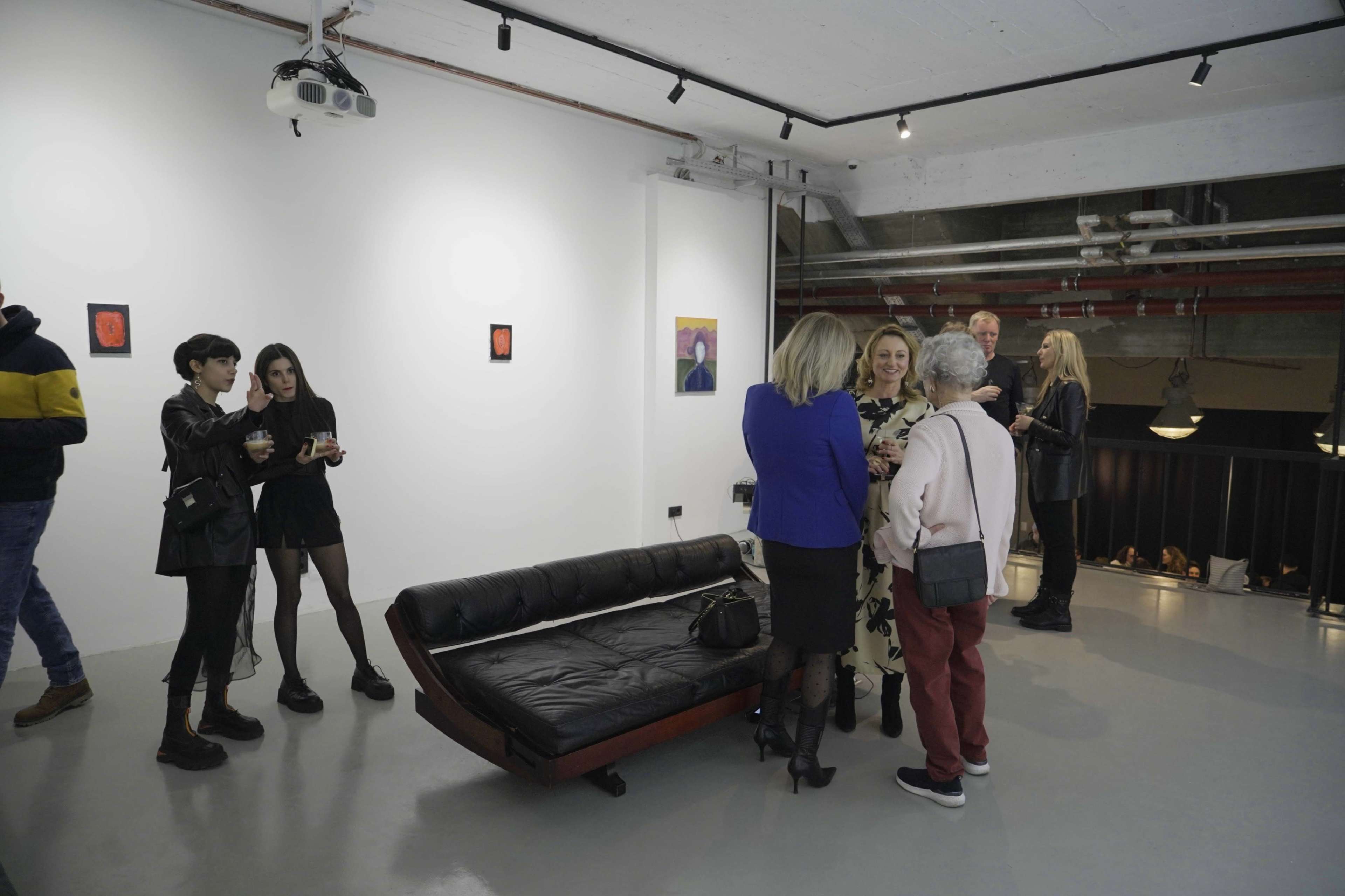 A group of people converse in an art gallery, with several artworks displayed on the white walls and a black couch positioned in the center.