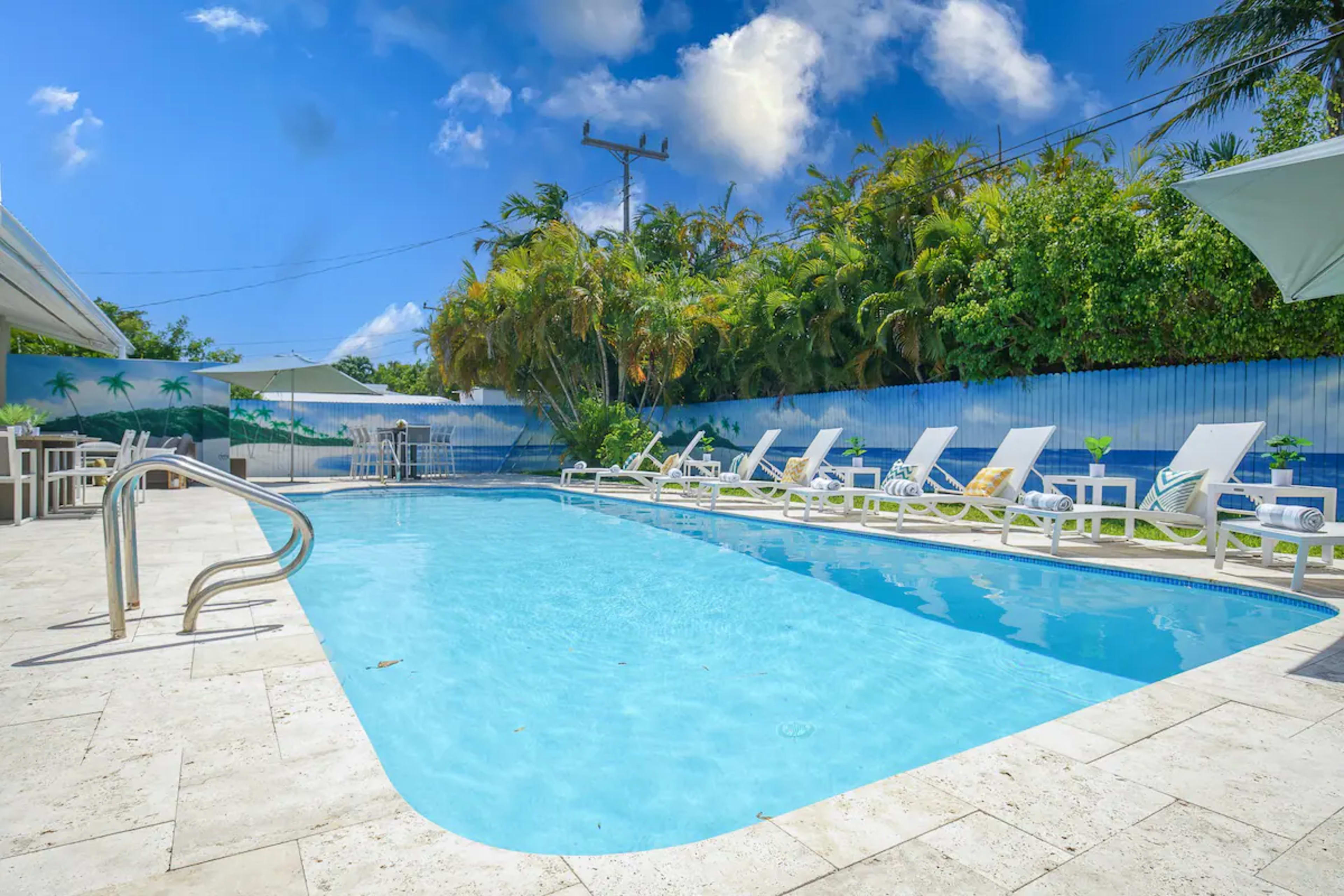 A clear swimming pool surrounded by lounge chairs and tropical plants under a blue sky.