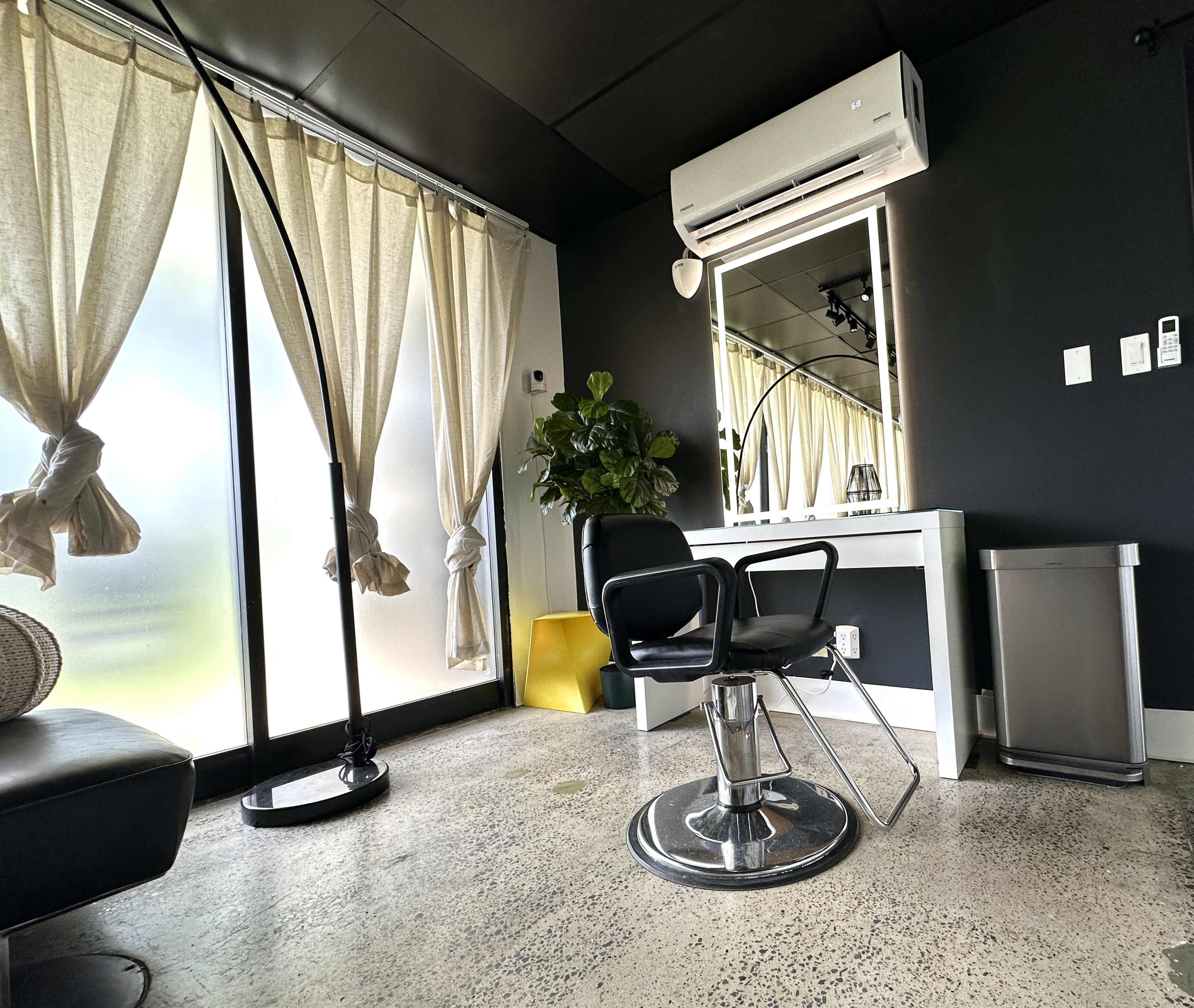 The image shows a modern salon space featuring a styling chair, a large mirror, a potted plant, and large windows with sheer curtains.