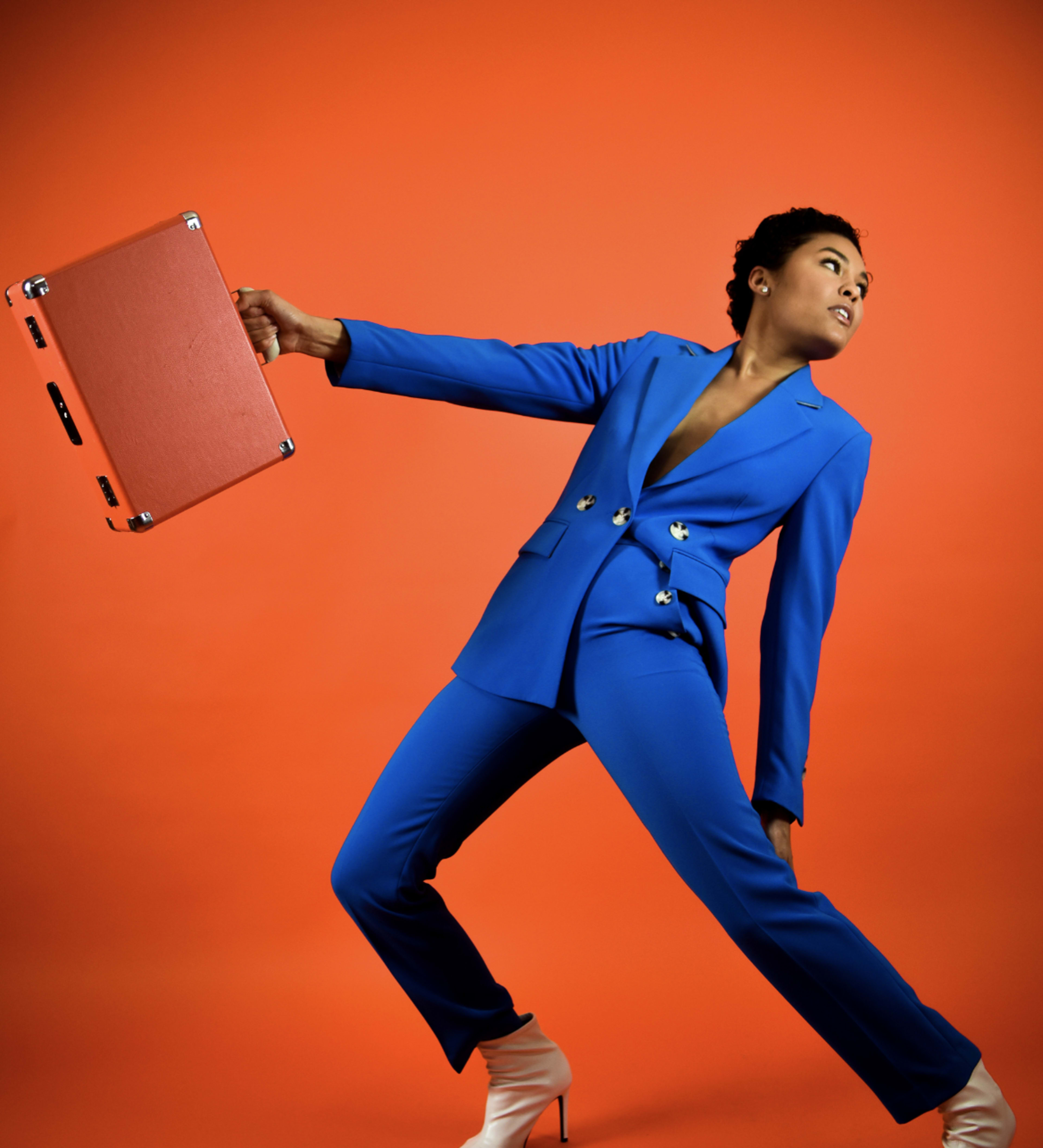 A person in a blue suit poses dramatically against an orange background while holding a red briefcase.
