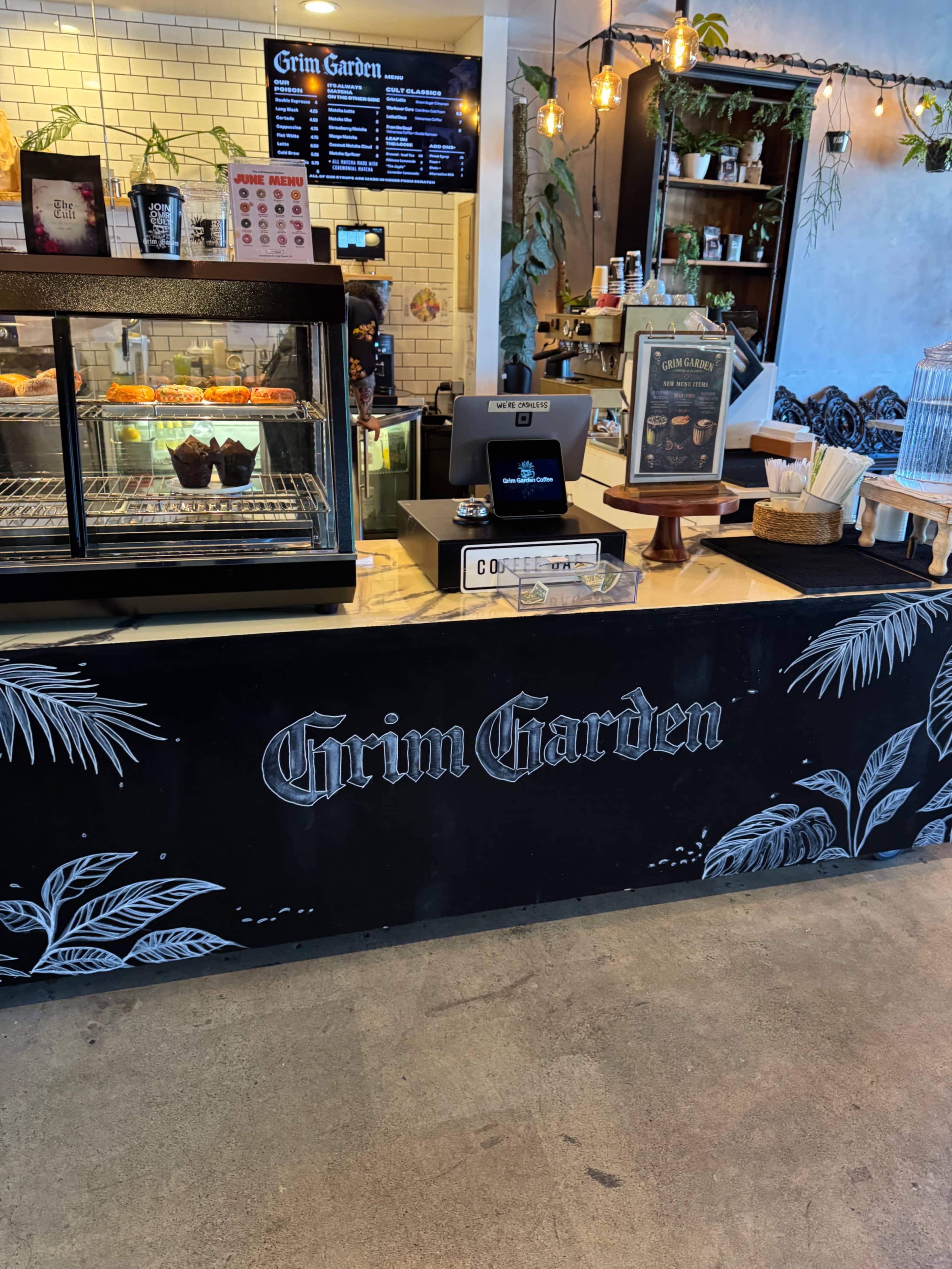 The image shows a café counter with a menu board, a display case for pastries, and indoor plants decorating the space.