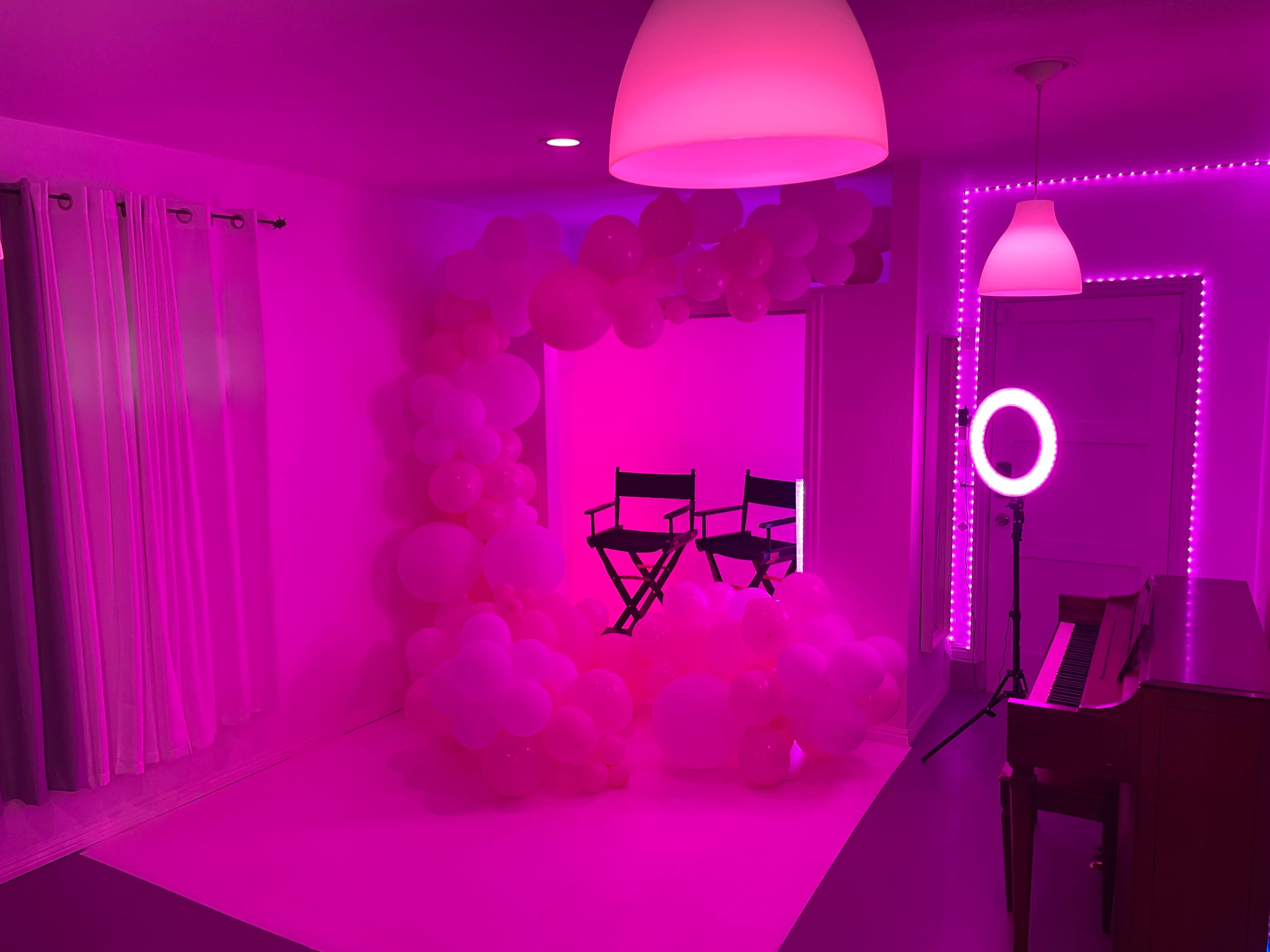 A brightly lit room decorated with pink balloons, featuring two director's chairs, a ring light, and a piano.