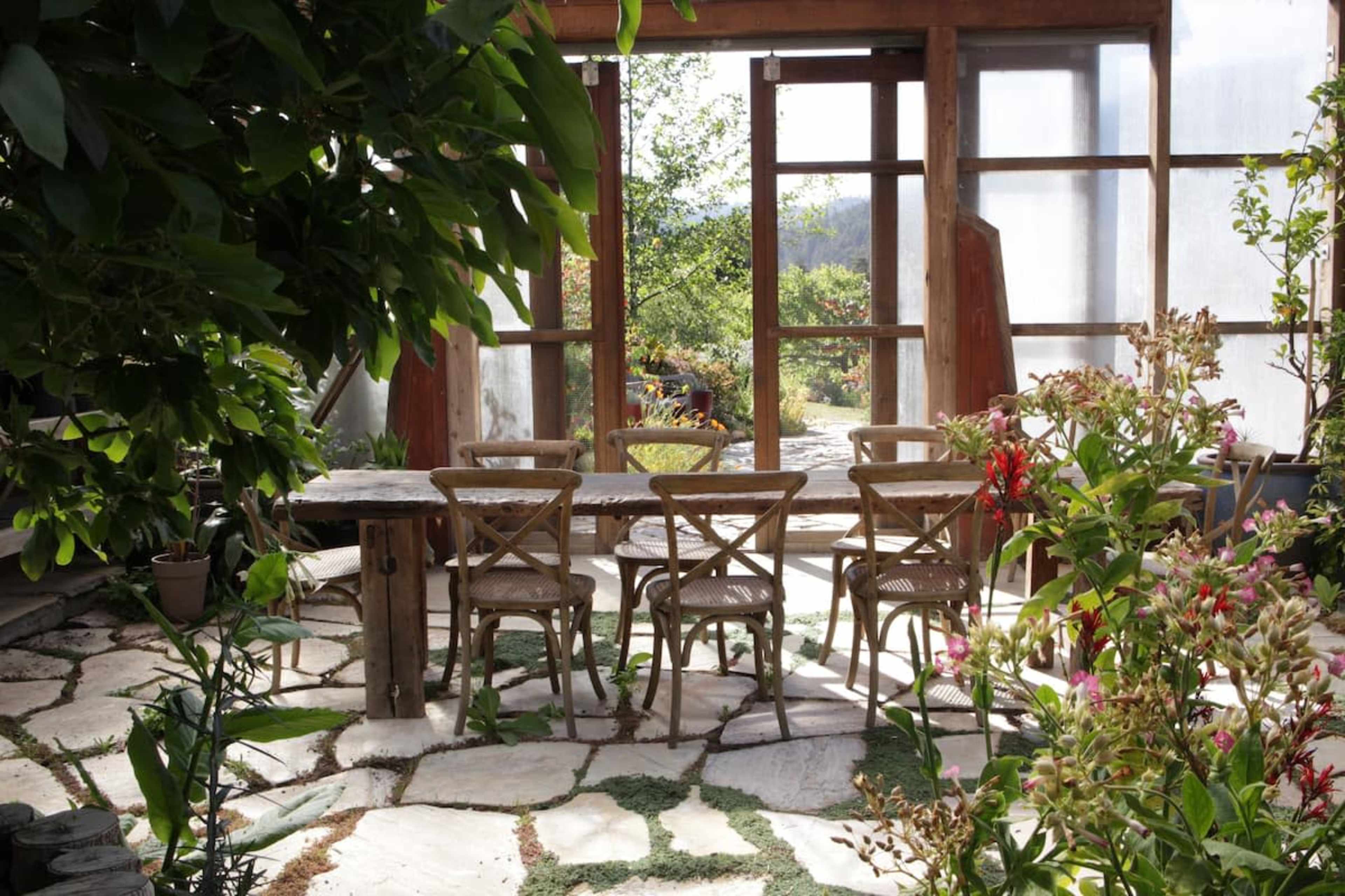 A cozy outdoor seating area with a wooden table and chairs surrounded by greenery and flowers, with large windows letting in natural light.