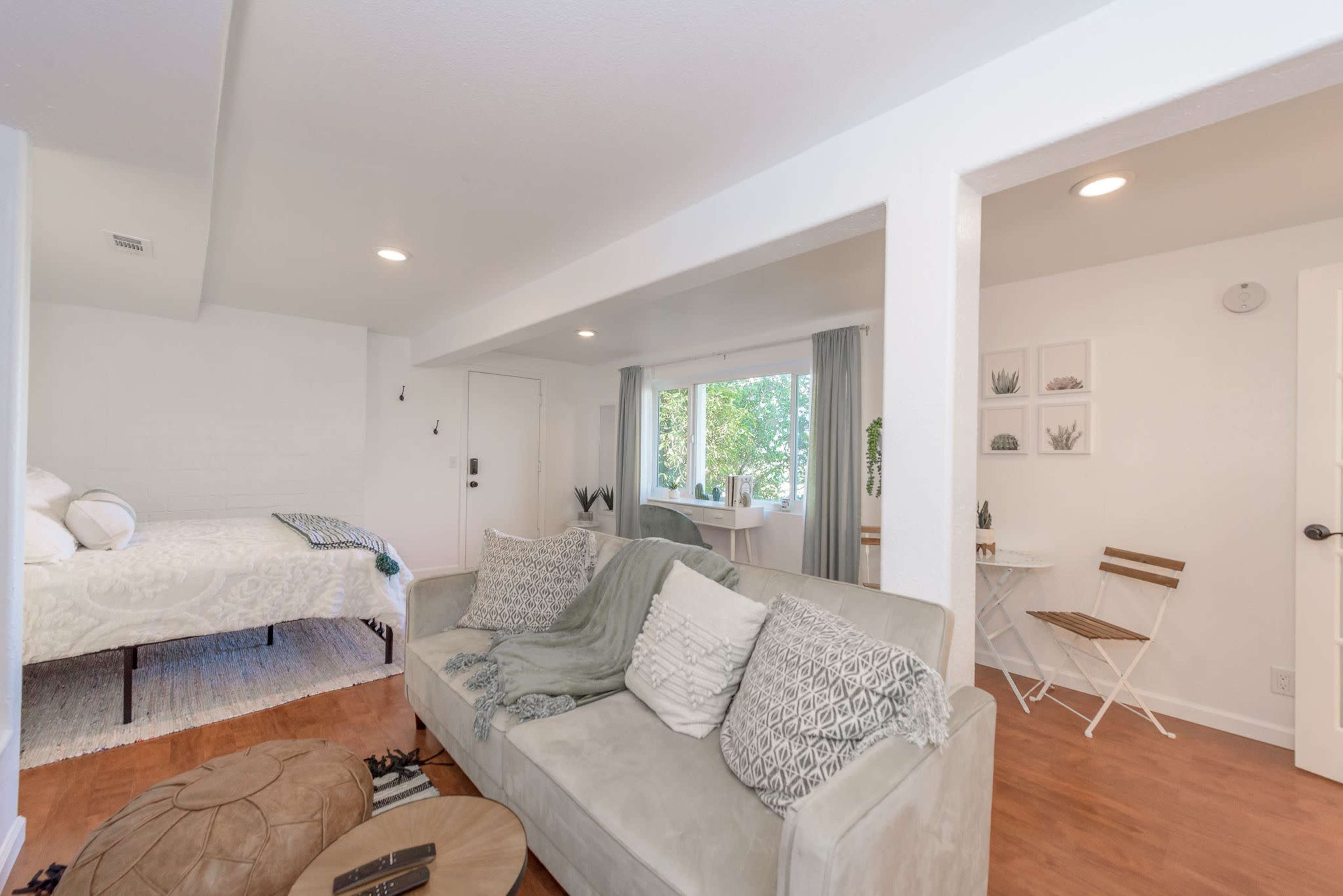 A well-lit, modern living space features a seating area with a sofa, a small dining table, and a bed visible in the background.
