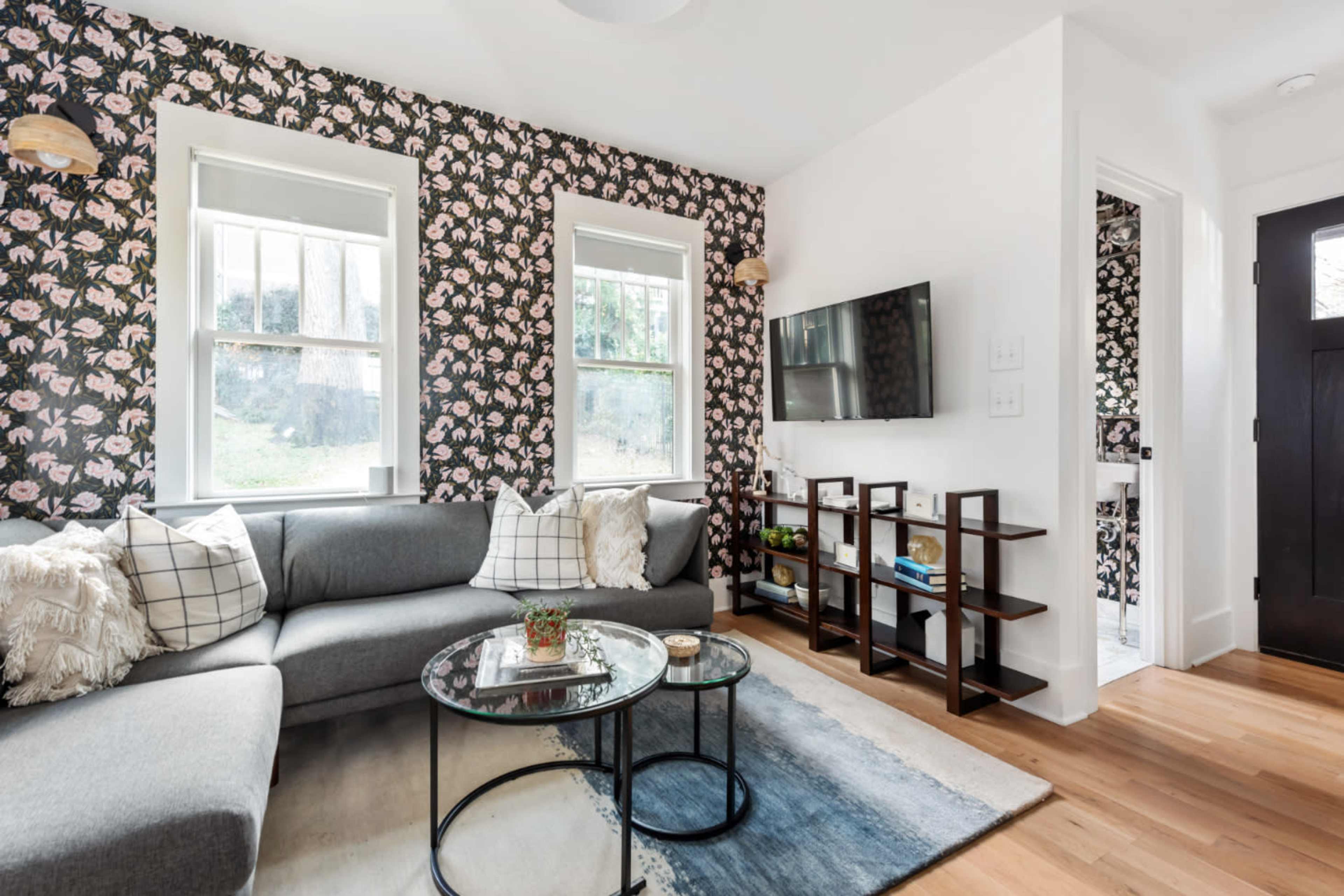 A modern living room featuring a gray sectional sofa, a glass coffee table, floral wallpaper, and a wall-mounted television.