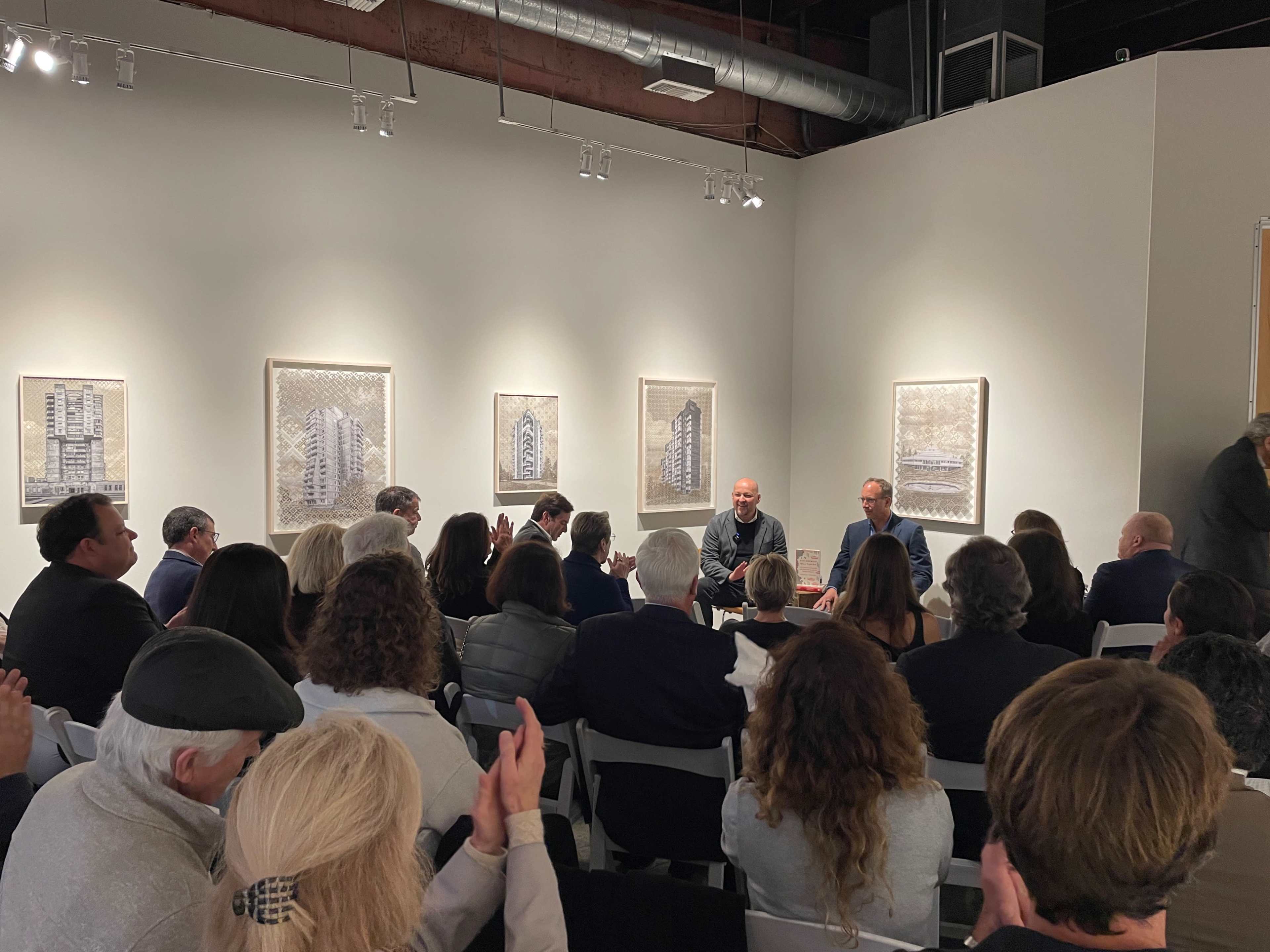 A crowded art gallery features three speakers seated at a table, with several framed artworks displayed on the walls behind them.